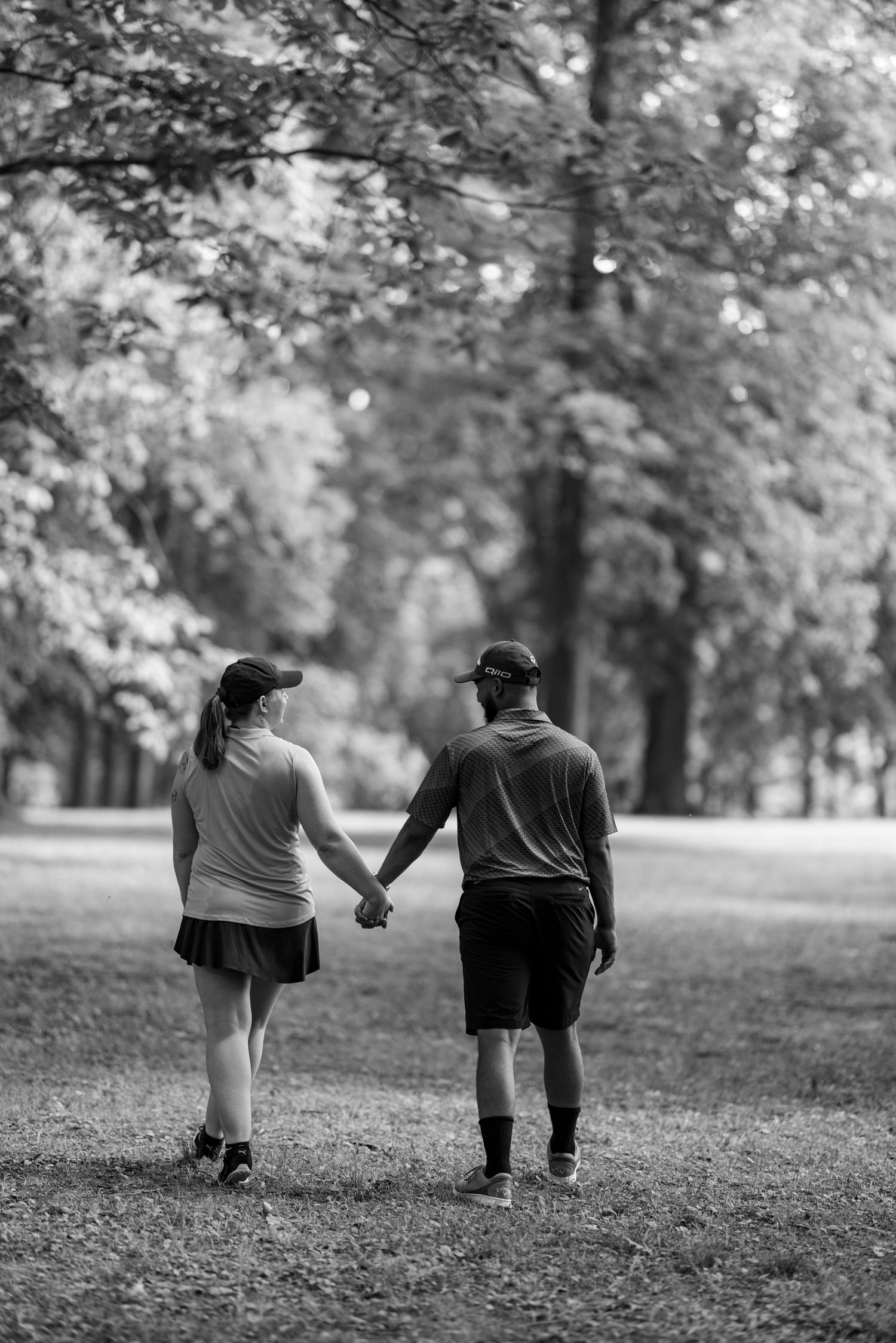 A couple holds hands while walking through a lush green park in black and white.