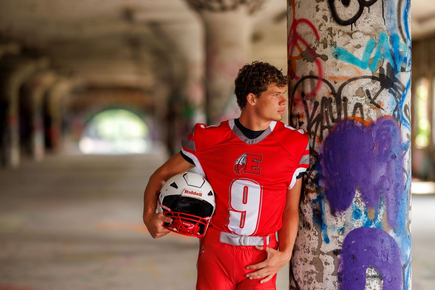 Football player in red number 9 jersey leaning against a graffiti-covered pillar under a bridge.
