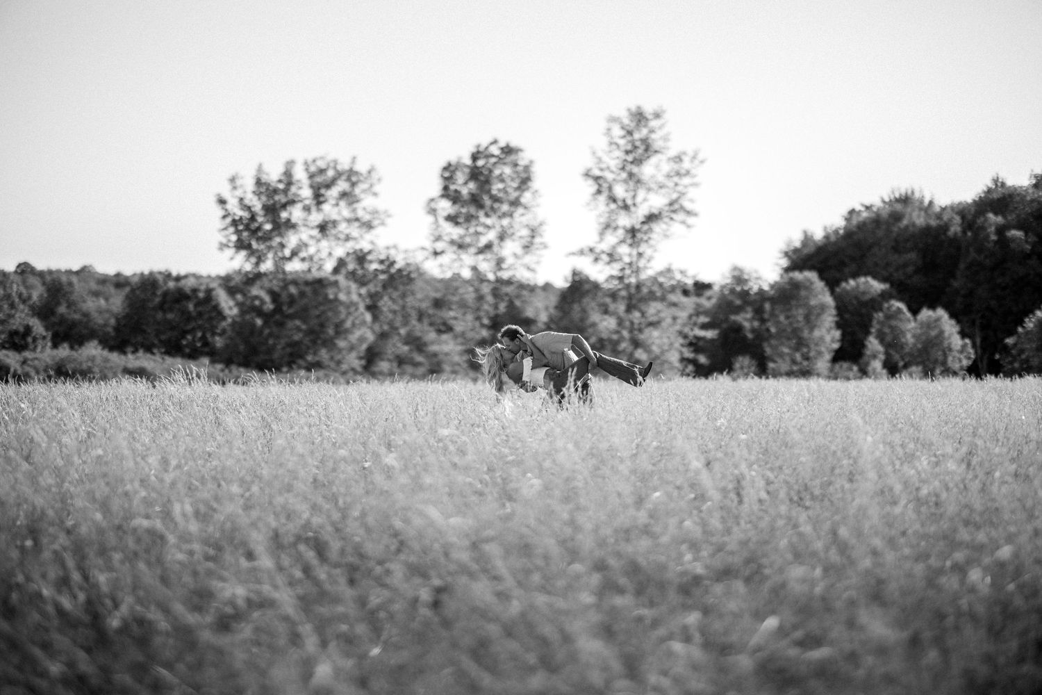 Black and white photo of couple walking through tall grass field with trees behind.