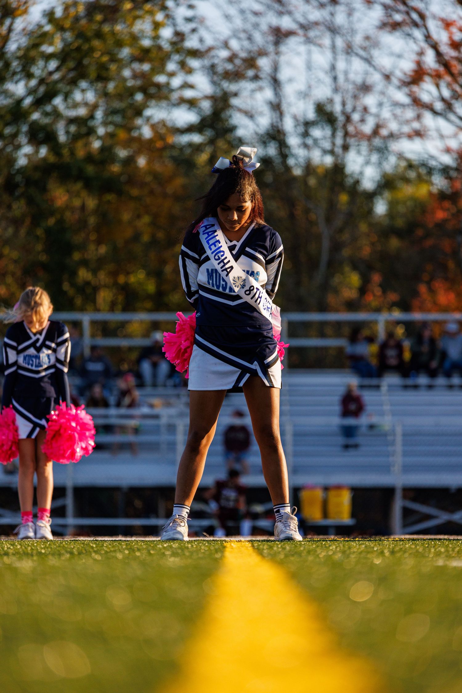 High school cheerleader in navy uniform with sash performs on a sunny autumn football field sideline.