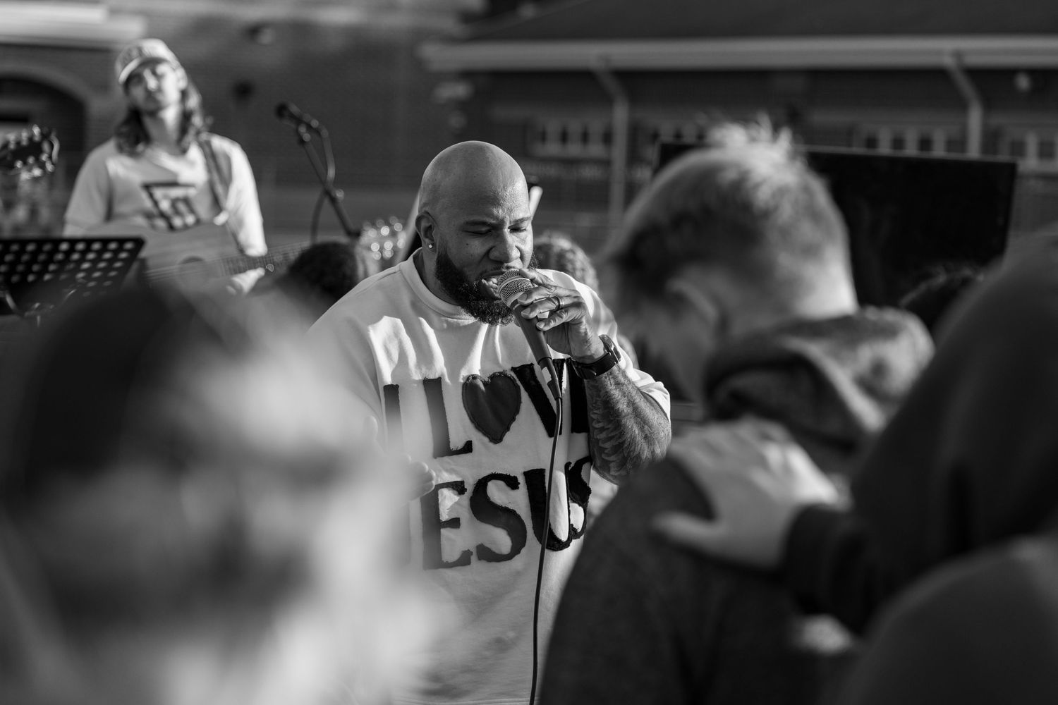 Man in I Love Jesus shirt prays with microphone at an outdoor Christian worship event.
