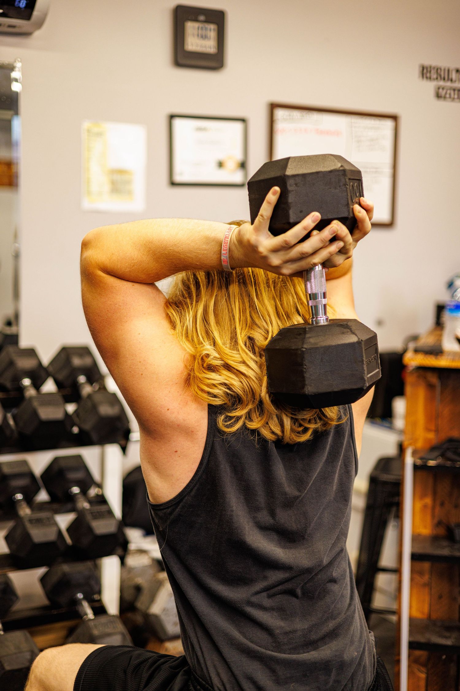 Woman performs overhead tricep extension with heavy dumbbell in gym with dumbbell rack in background.