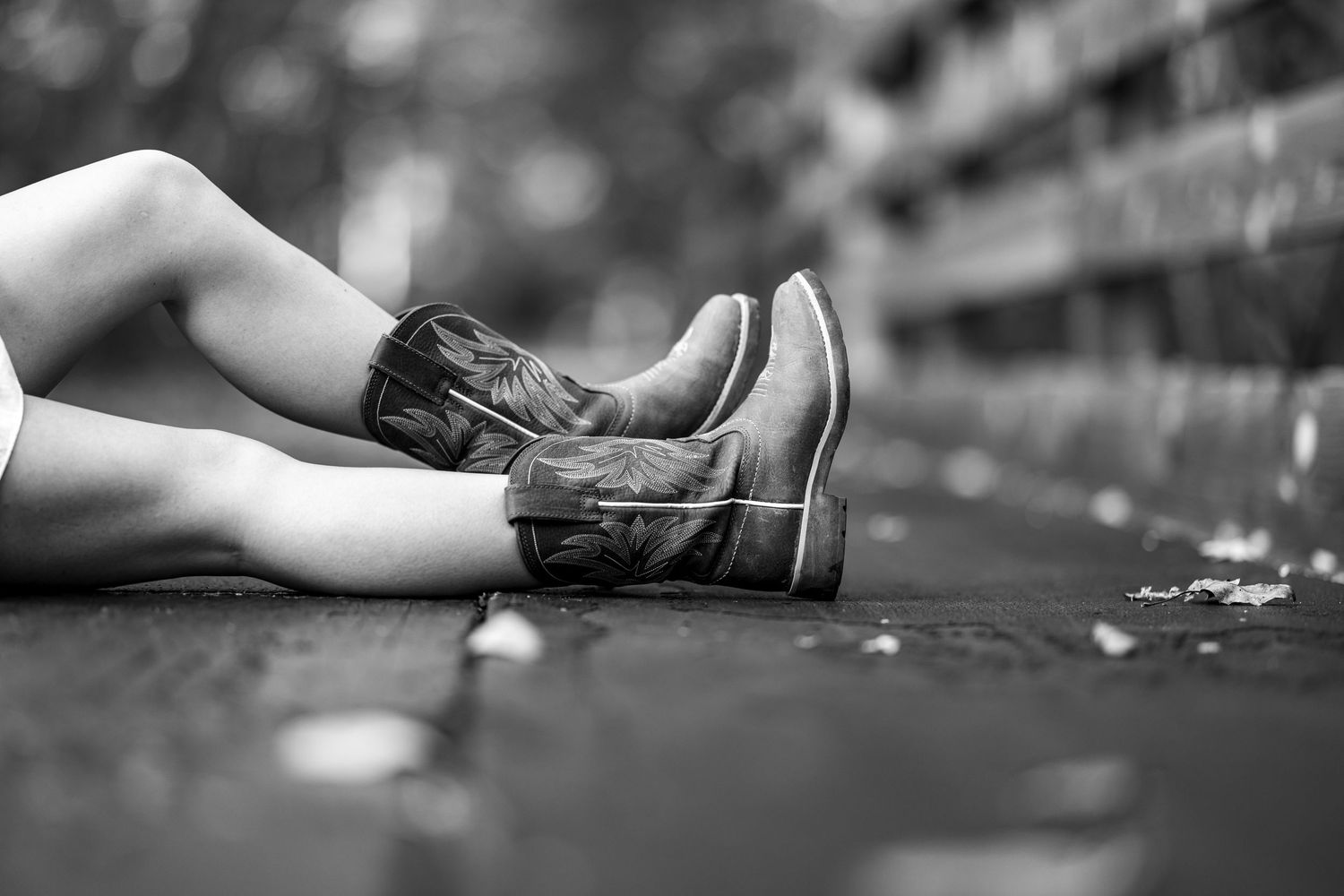A black and white close-up of a womans legs wearing cowboy boots sitting on a wooden bridge.