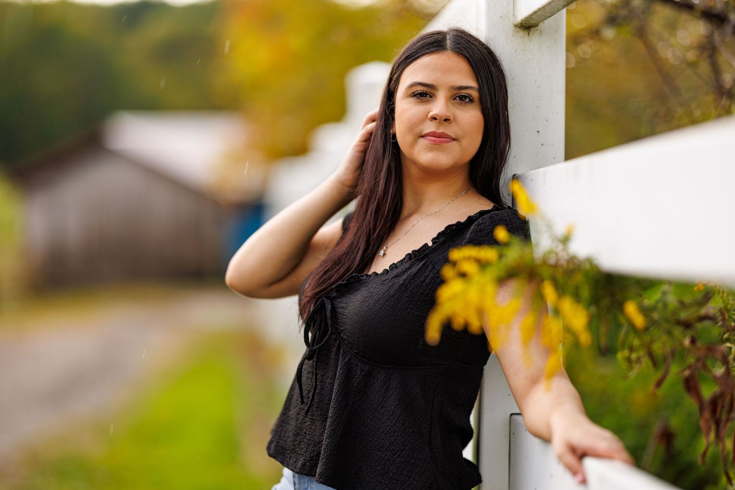 Brunette teen posing by white fence with yellow wildflowers in autumn setting.