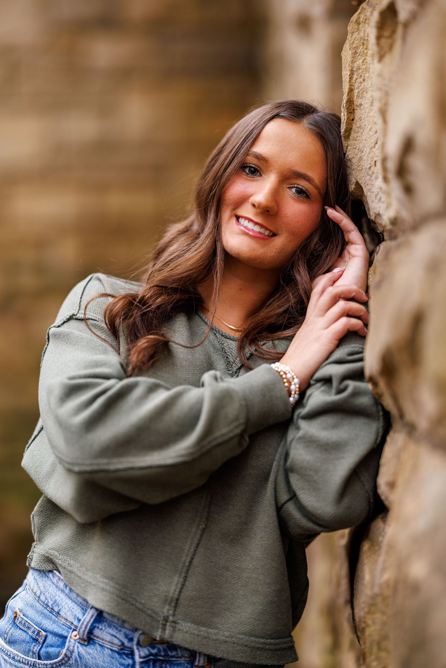 Smiling teen girl in olive sweatshirt and jeans leaning against rustic stone wall.