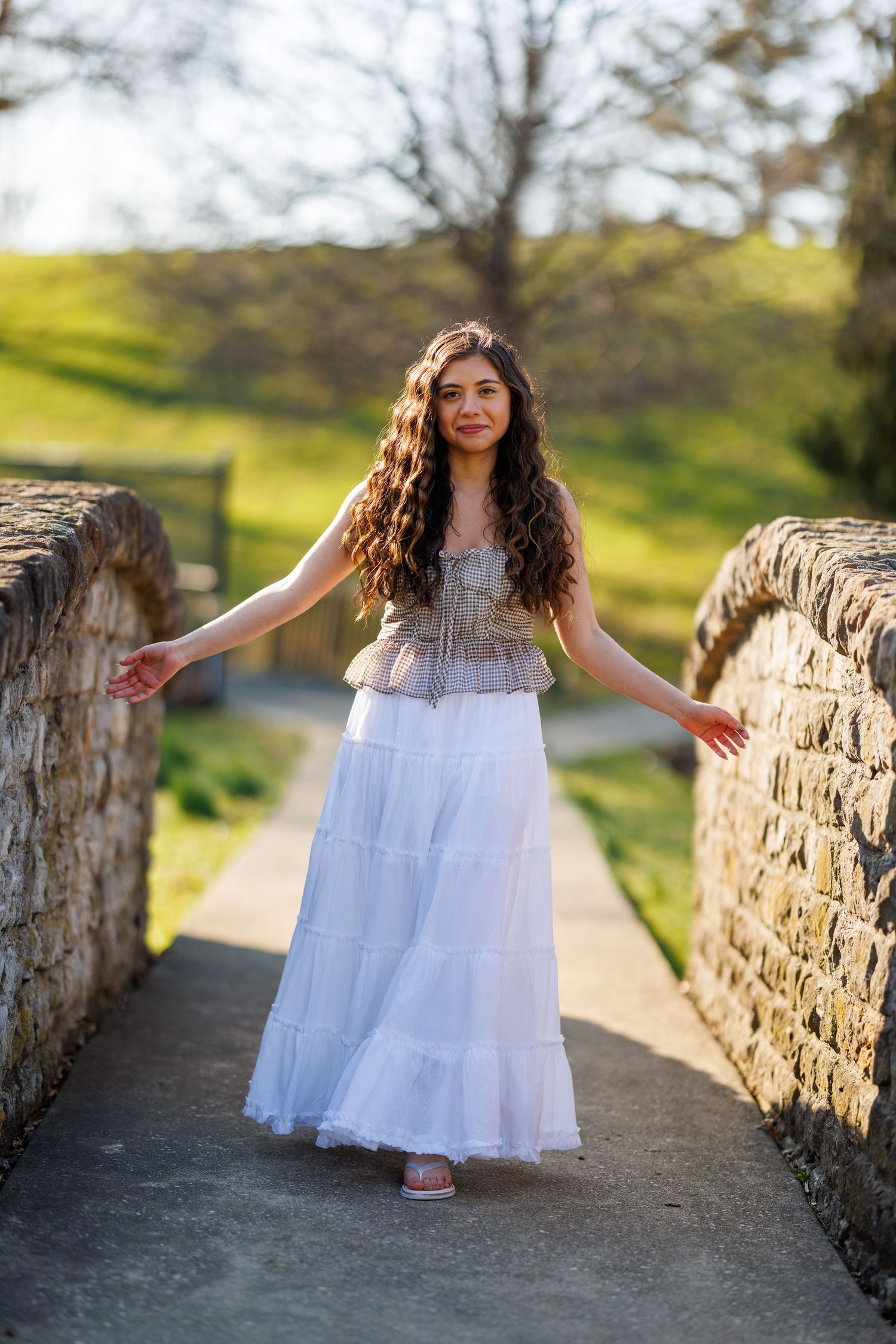 Young woman in white skirt and corset top posing on a rustic stone bridge in a park.