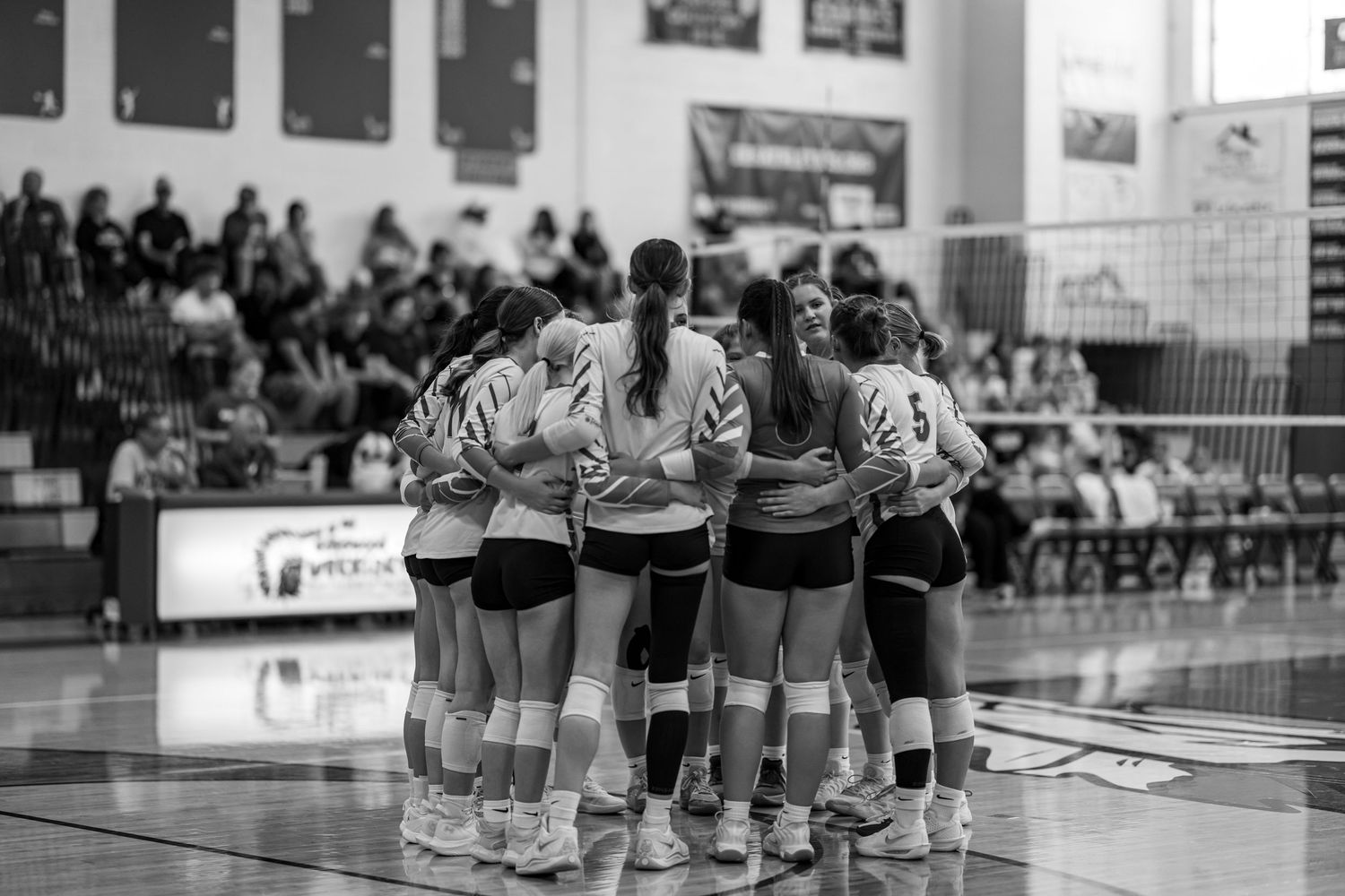 Black and white photo of a girls volleyball team huddling together on the court before a match.