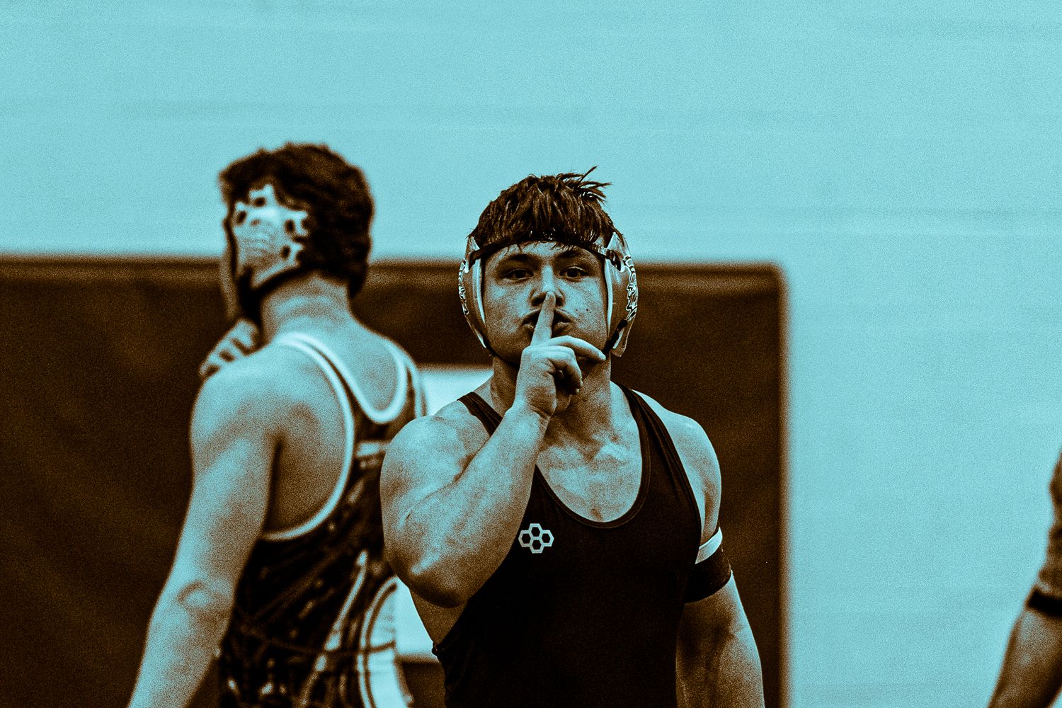 A wrestler in a black singlet makes a silence gesture with his finger to his lips during a match.