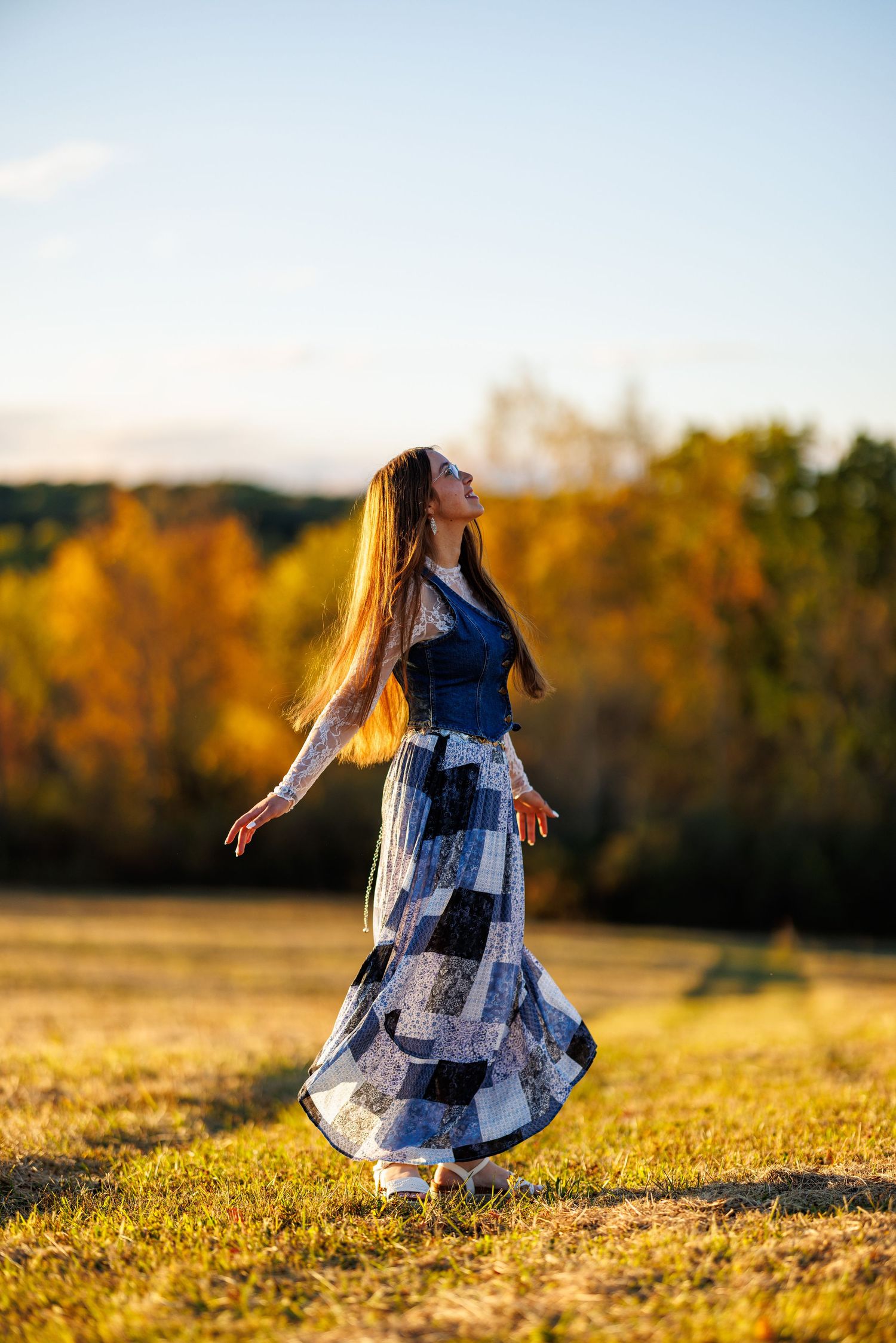 Young woman in blue patchwork maxi dress twirling in open field with colorful autumn foliage background.