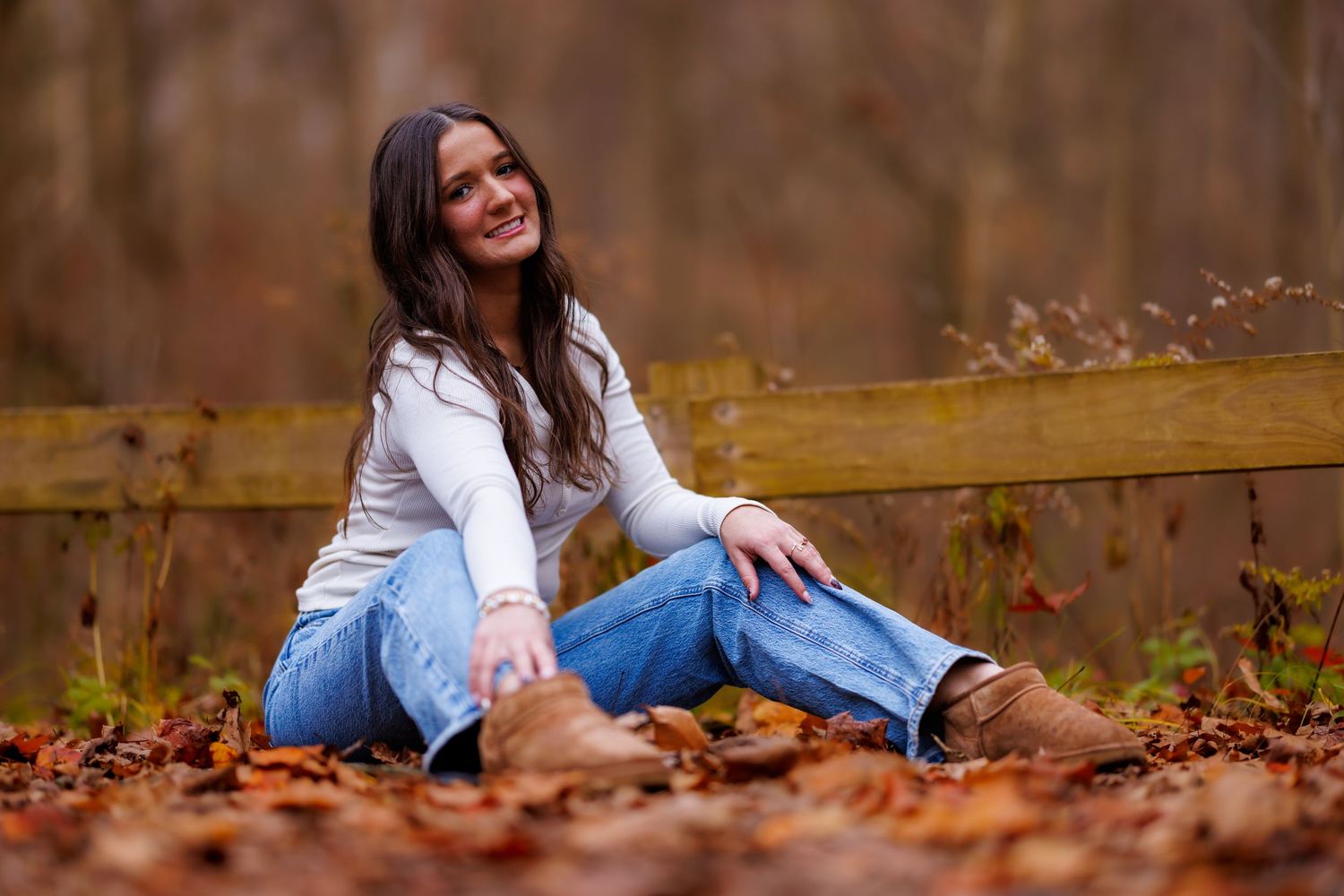 Smiling teen girl in white top and jeans sitting on fallen leaves by wooden fence.