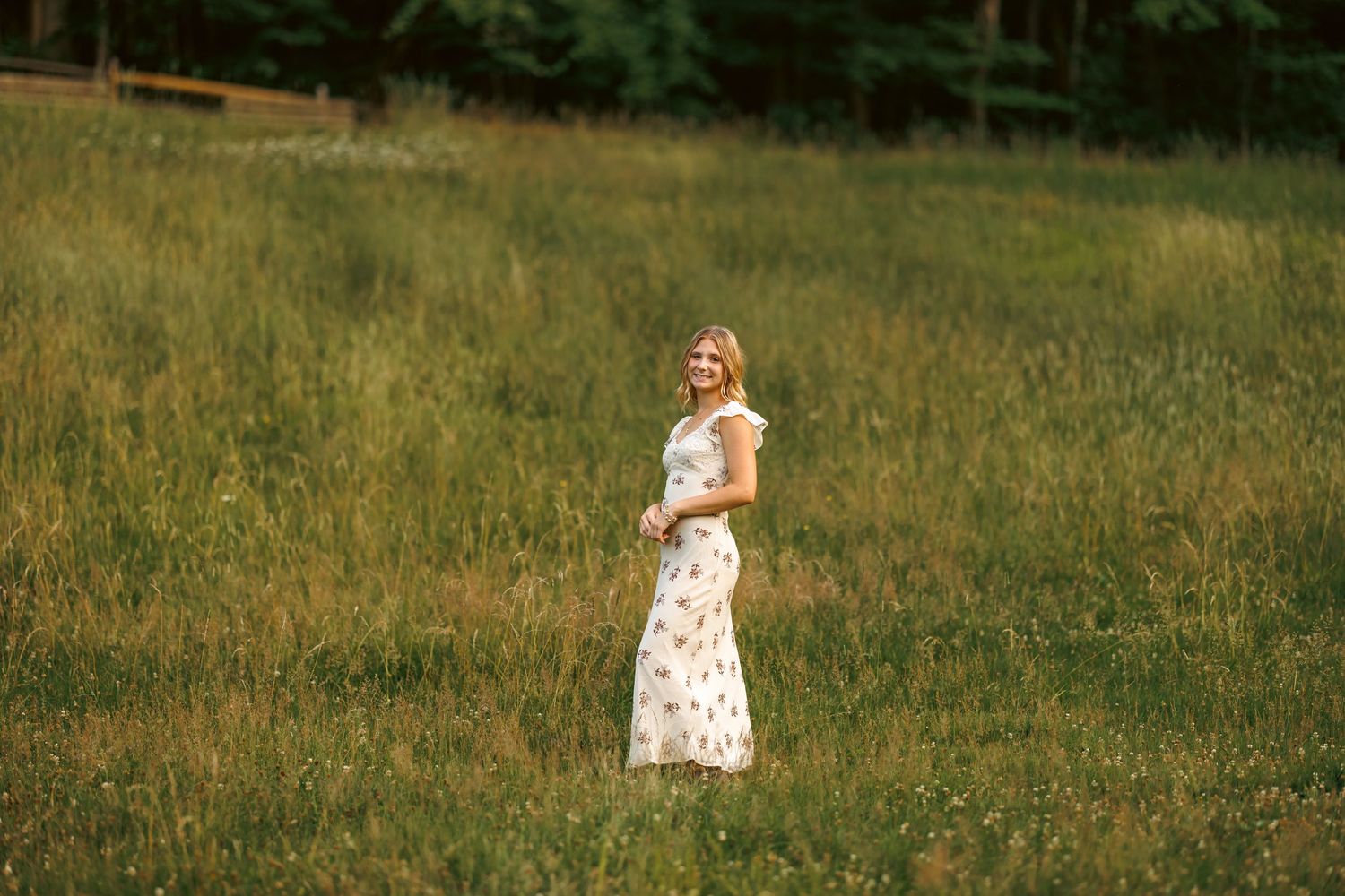 Blonde woman in a white floral maxi dress standing in a sunlit green meadow with trees behind her.
