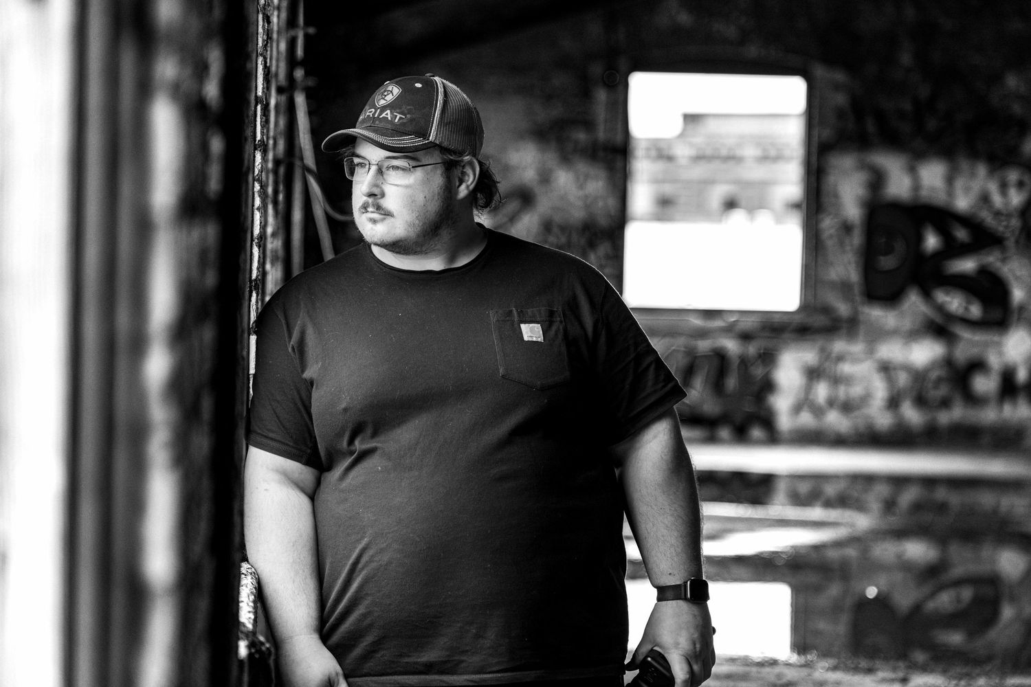 Black and white portrait of man in cap and t-shirt standing in abandoned graffiti building.