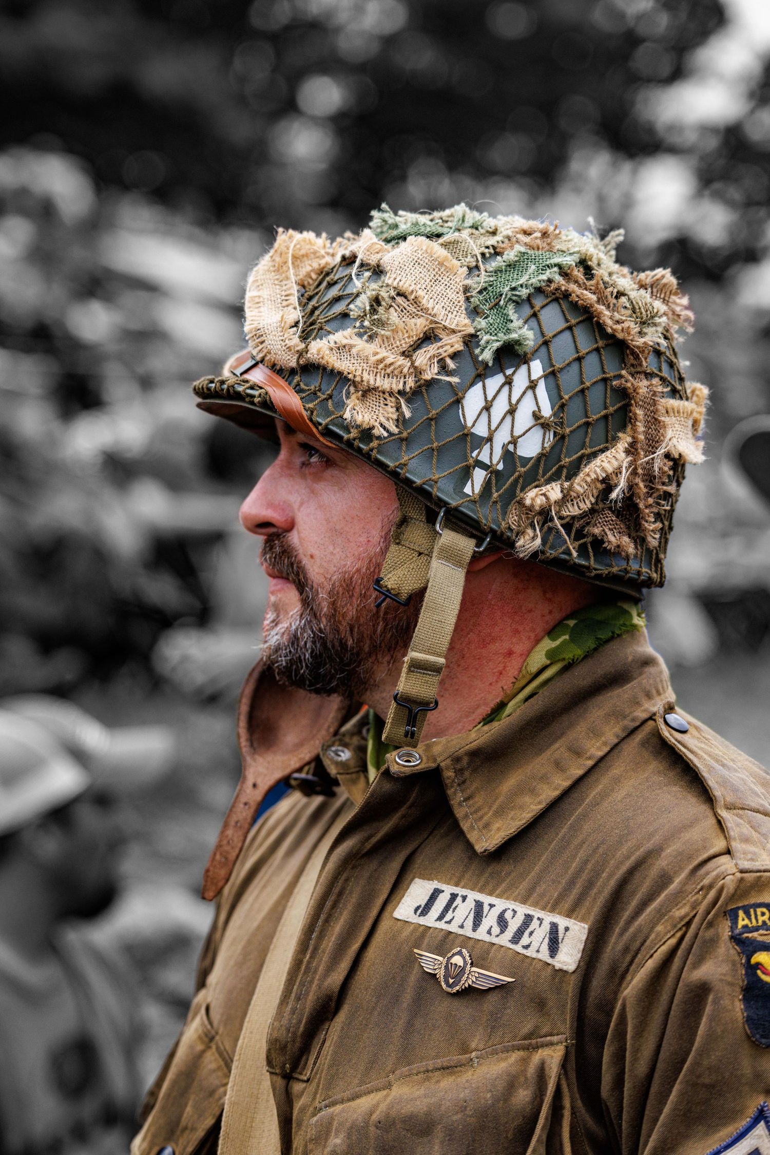 Bearded soldier in WWII US Airborne uniform with camouflaged helmet at military reenactment event.