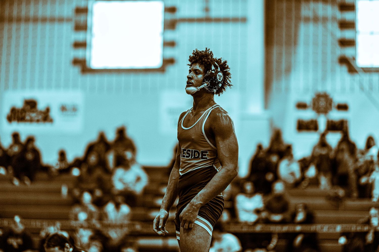 A muscular wrestler in a yellow singlet stands confidently on the mat with a crowd in the background.