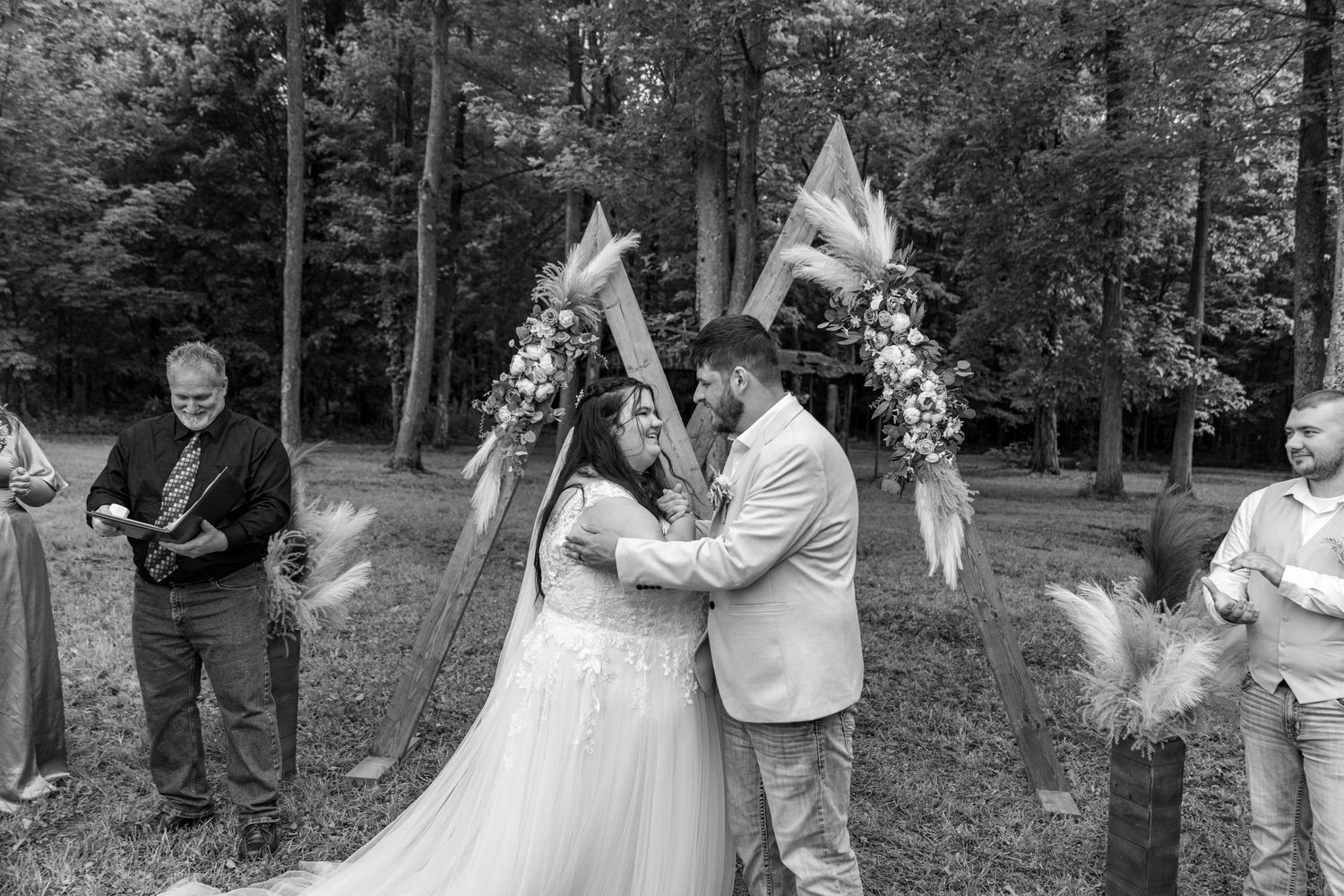 Bride and groom share their first kiss at an outdoor woodland wedding ceremony with guests celebrating.