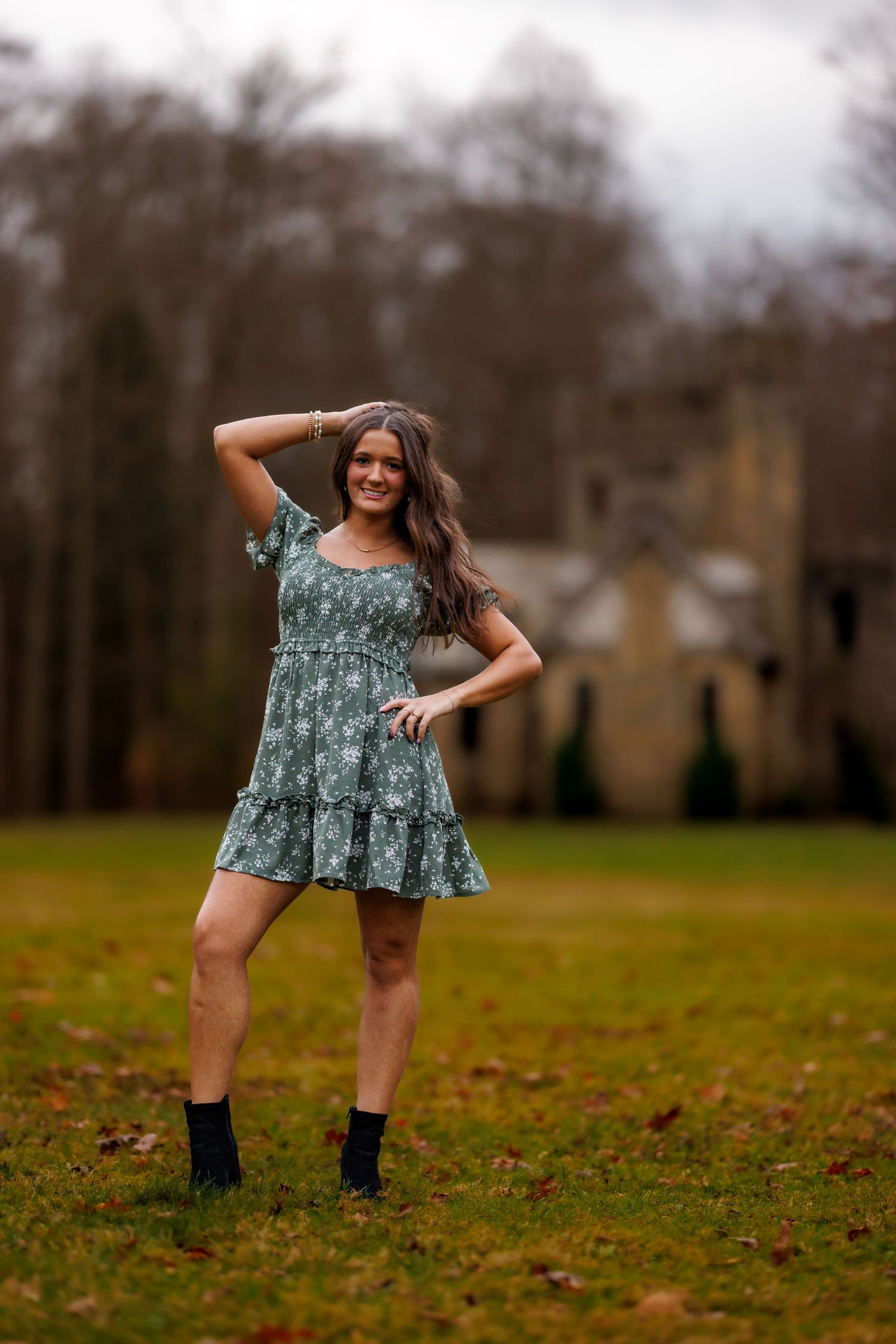 Young woman in green floral dress and boots poses in autumn field near historic building.