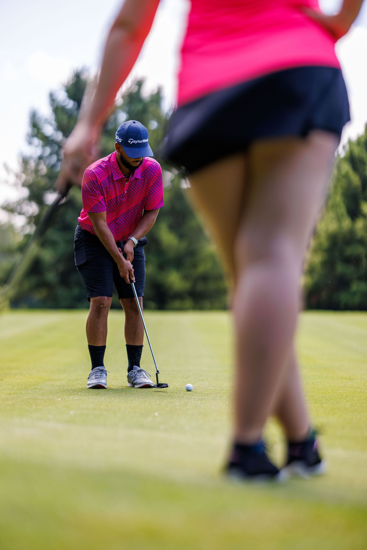 A golfer in a pink shirt focuses on a putt while another player watches from the foreground on a green course.