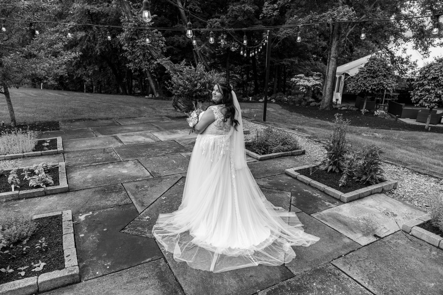 A bride in a flowing white gown and veil twirls gracefully in a garden courtyard in this black and white photo.