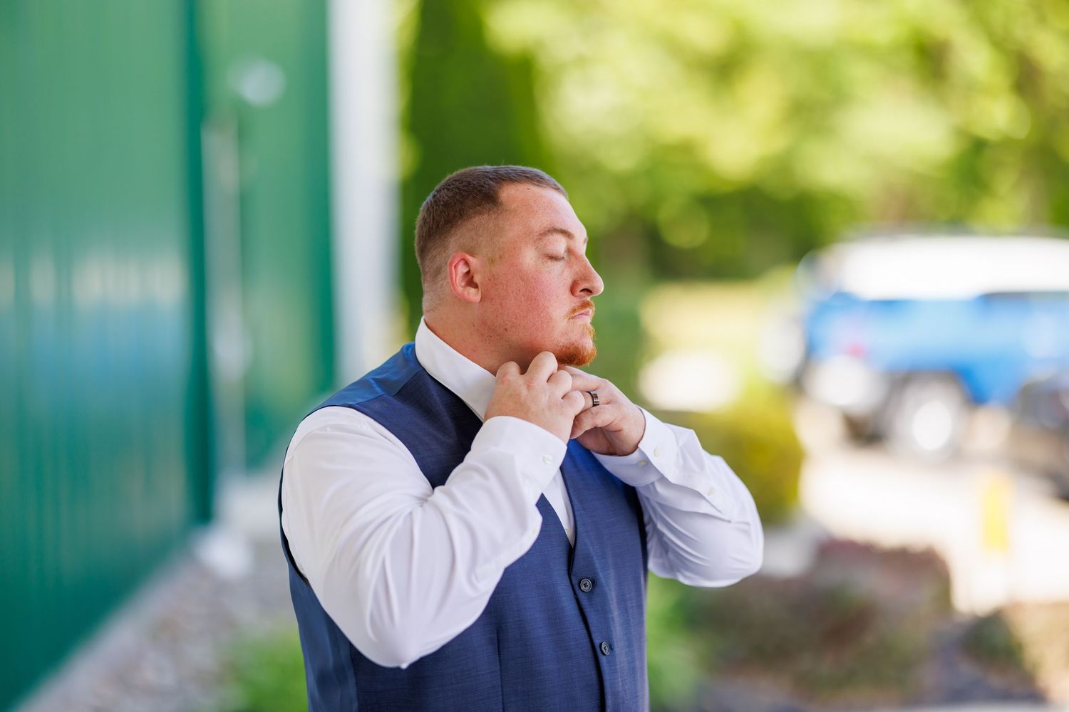 Groom in blue vest adjusting collar outdoors before wedding ceremony.