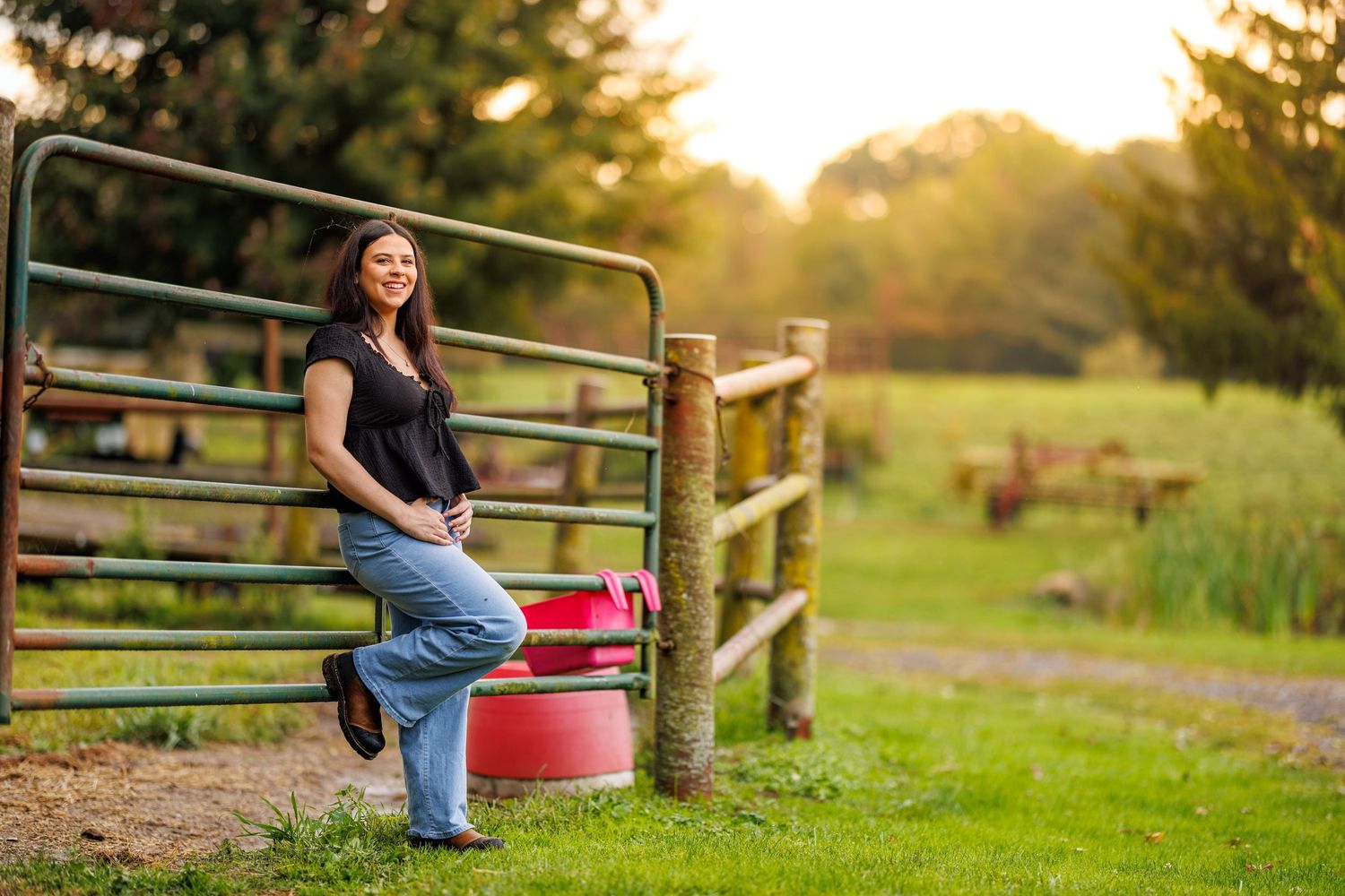 Woman leaning on farm gate smiling at sunset with green pasture background.