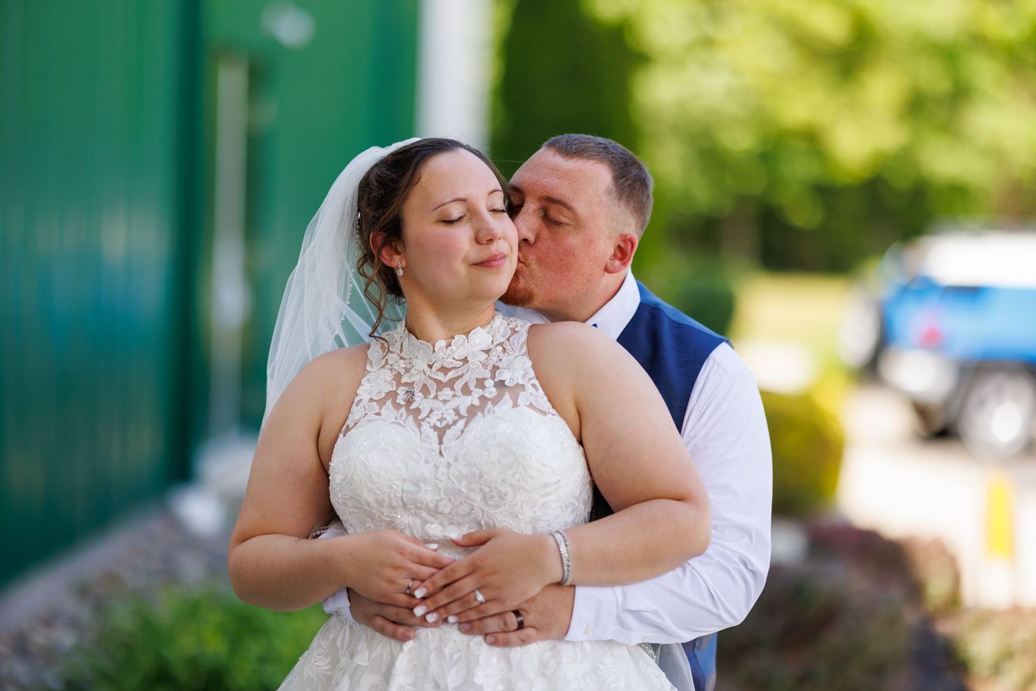 Groom kissing bride on cheek while embracing her outdoors in wedding attire.