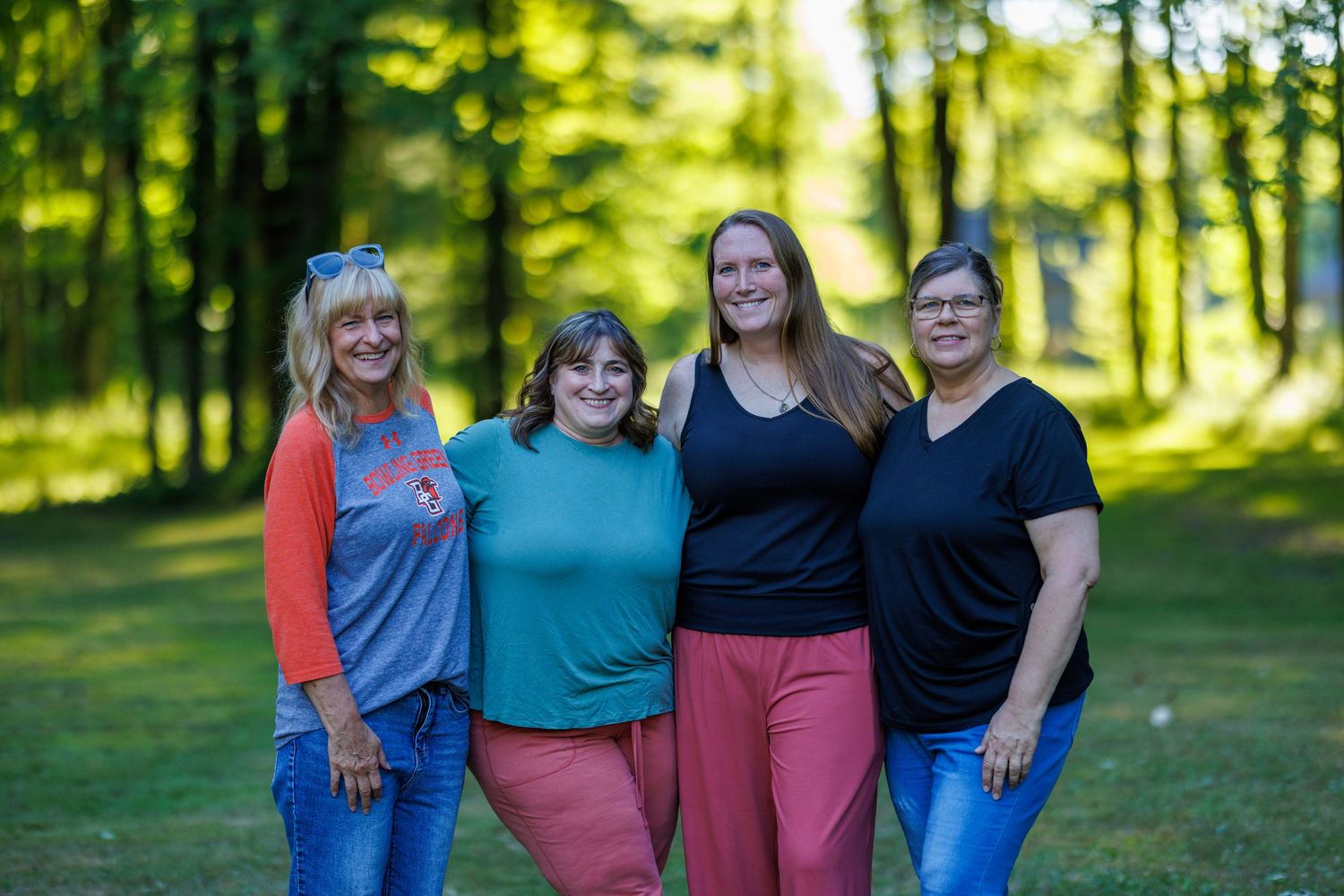 Four women smiling together outdoors in a green park setting on a sunny day.