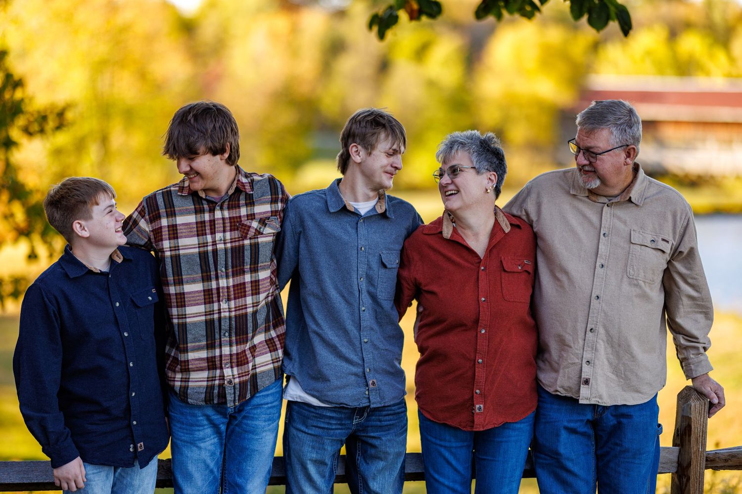 A joyful family of five laughing together outdoors in autumn, posing near a wooden fence with colorful fall foliage.