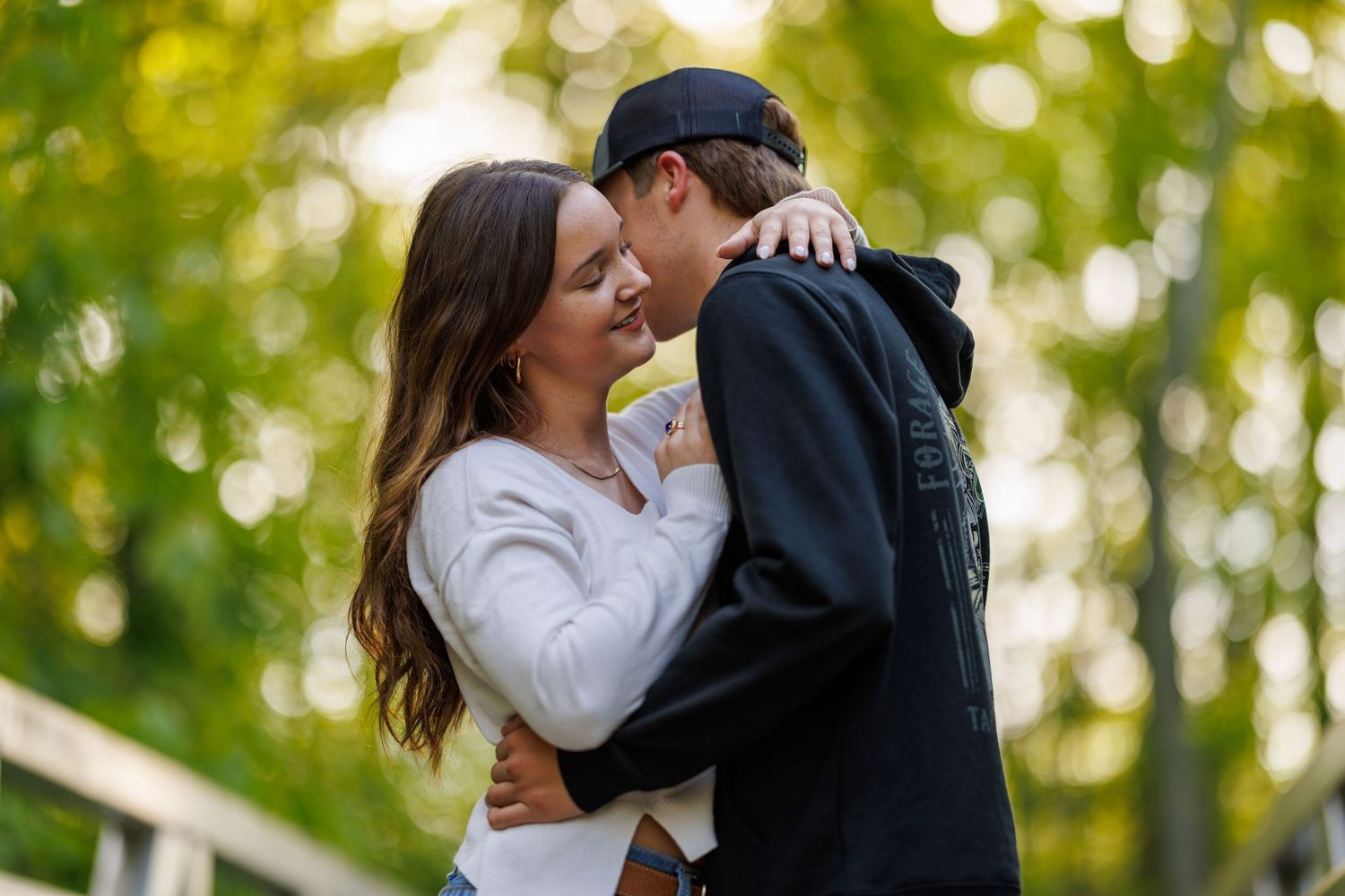 A couple sharing a tender embrace and almost kissing outdoors with lush green trees in the background.
