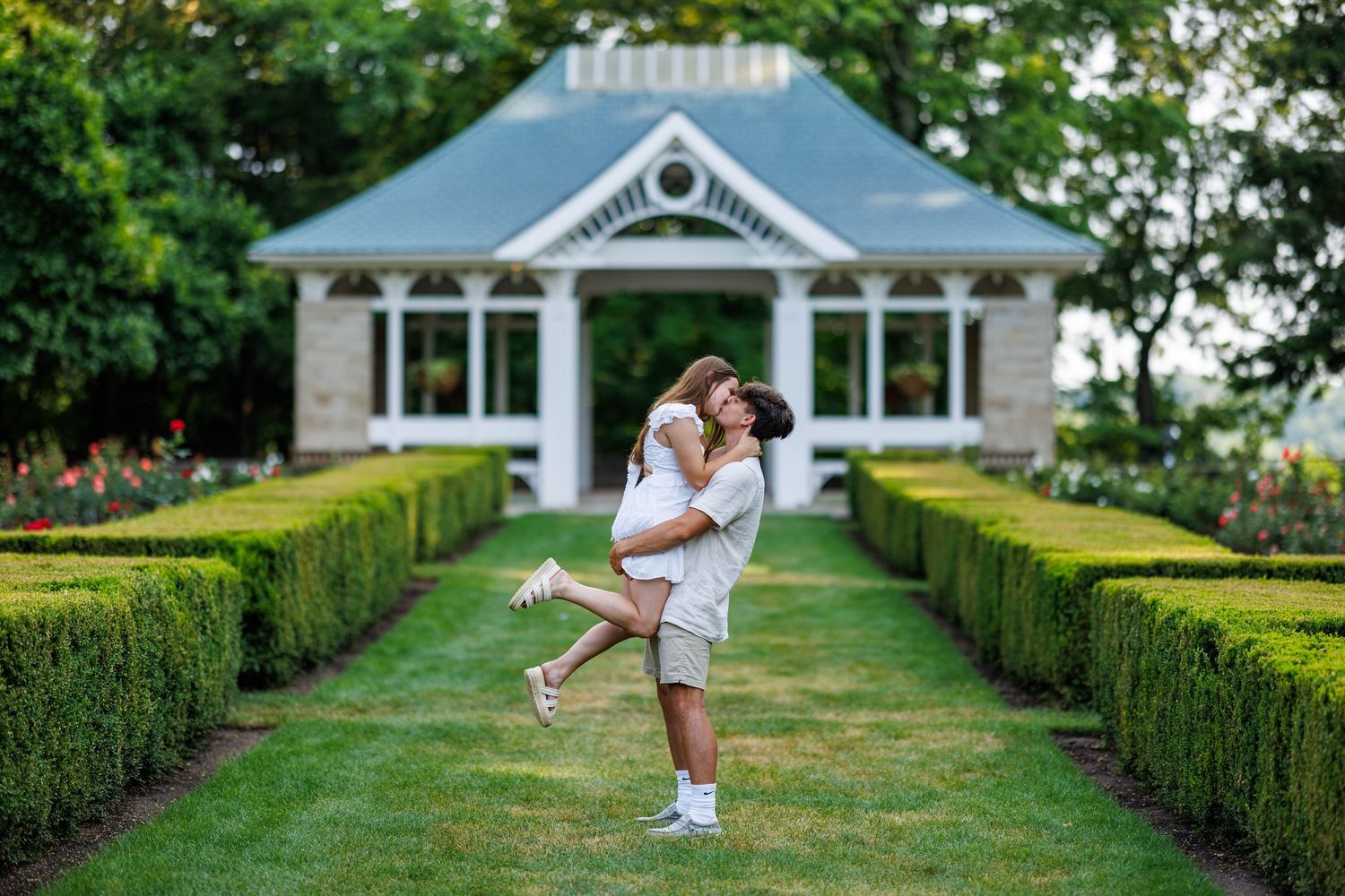 A man lifting his partner in a romantic garden with manicured hedges and a white gazebo backdrop.