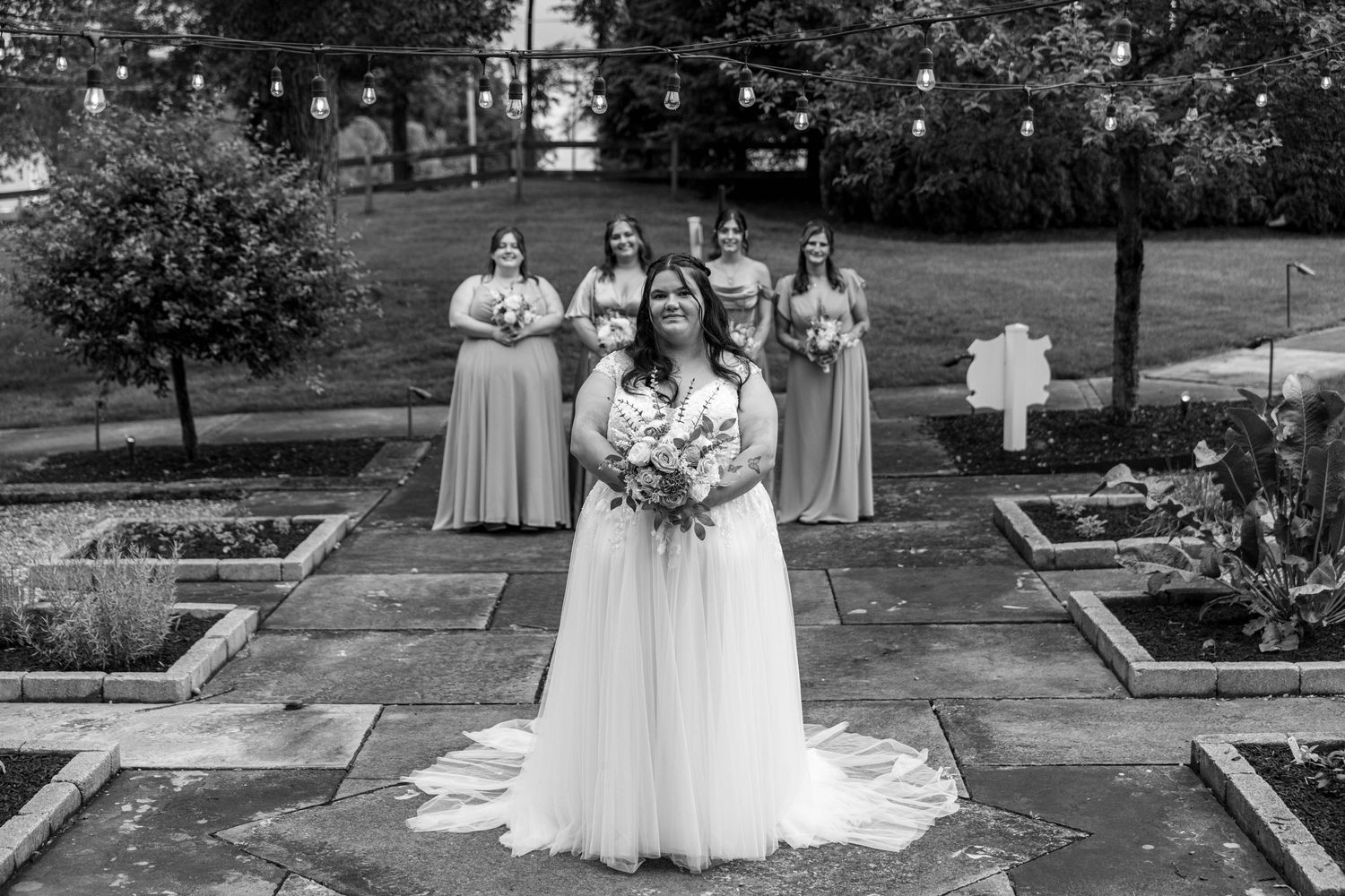 Bride poses with bridesmaids in garden setting, black and white wedding portrait with string lights.
