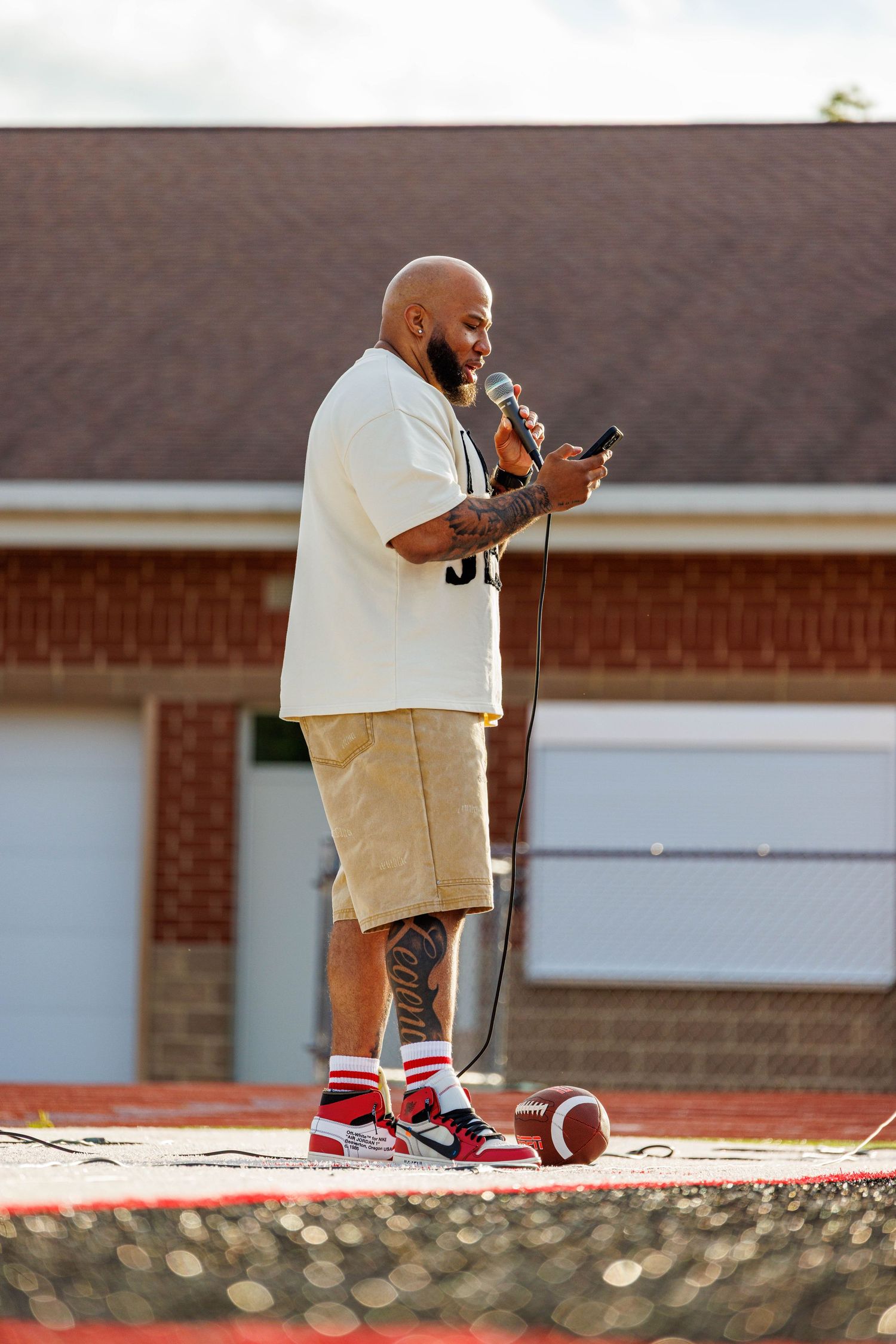 Tattooed bald man speaking into a microphone while holding a phone at an outdoor football field event.