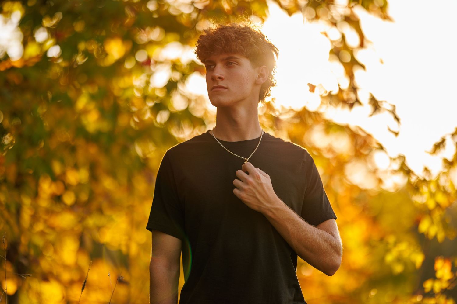 Young man in black t-shirt with gold chain poses among golden autumn foliage at sunset.