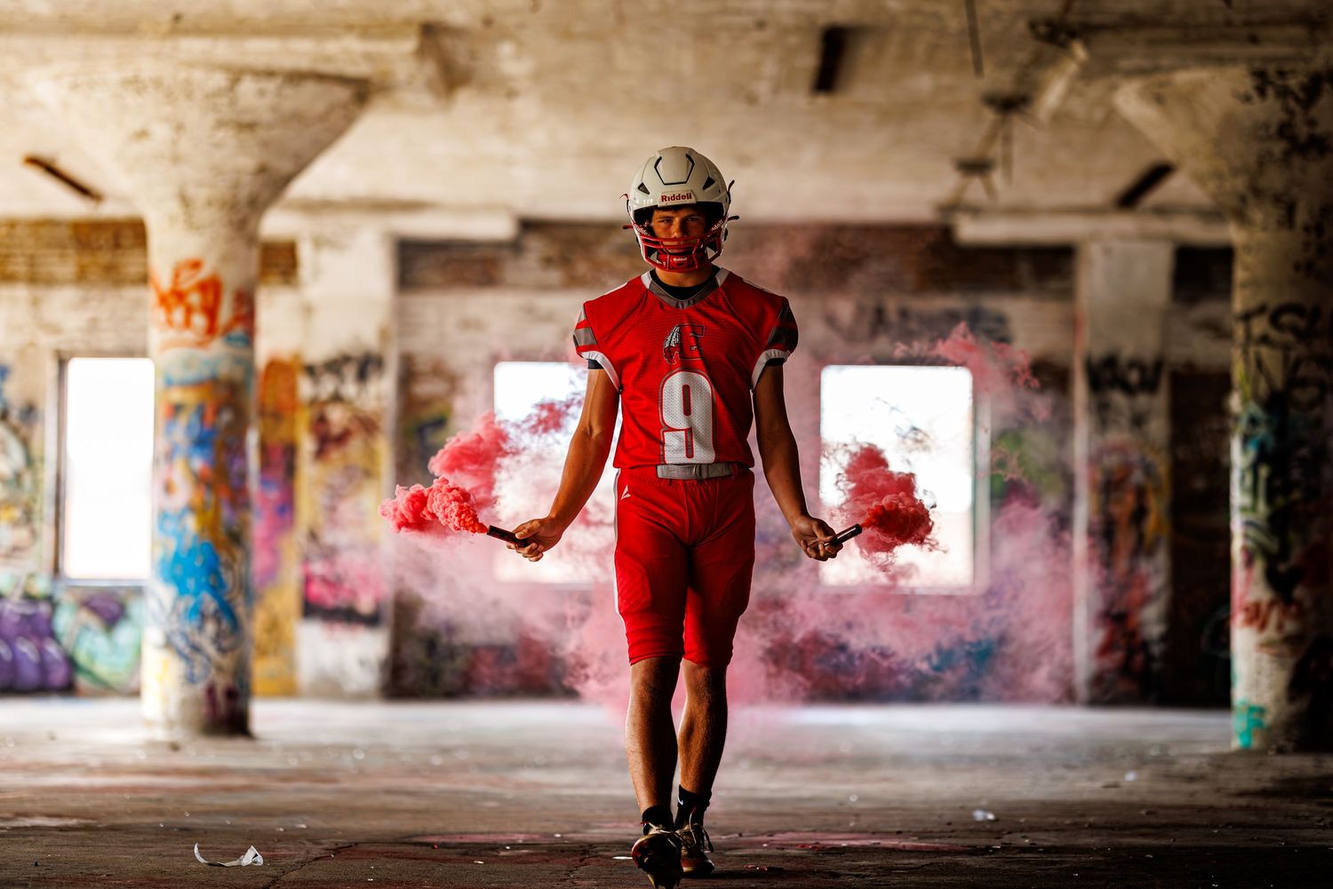 Football player in red uniform holding a red smoke grenade inside an abandoned graffiti-covered building.