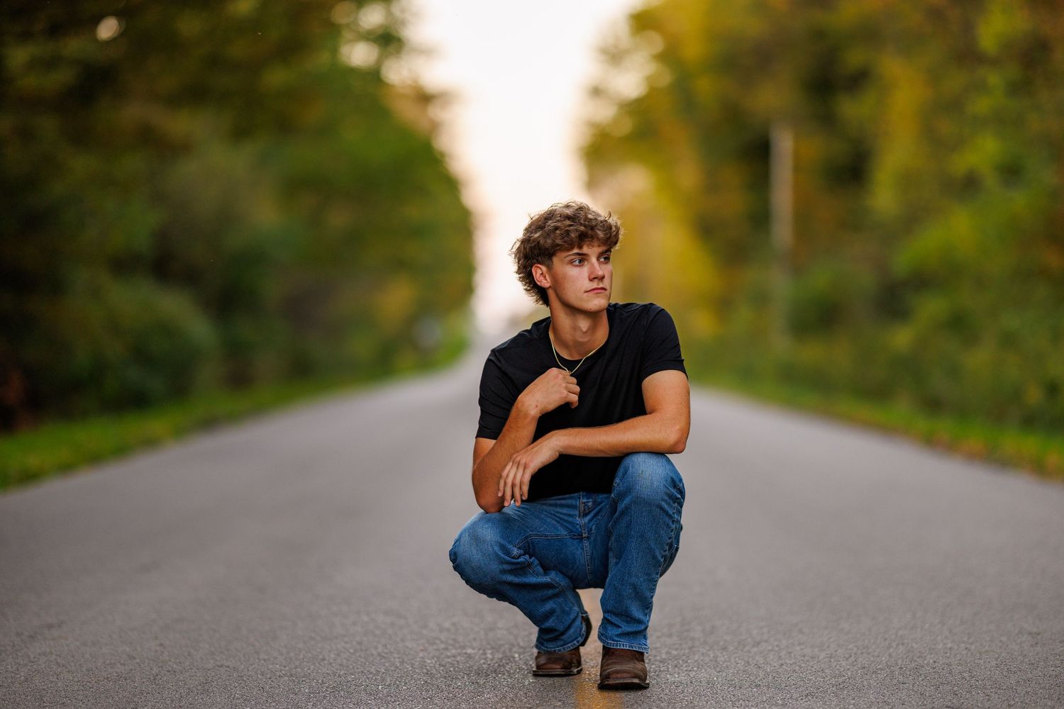 Young man in black t-shirt and jeans crouching on empty tree-lined road in autumn.
