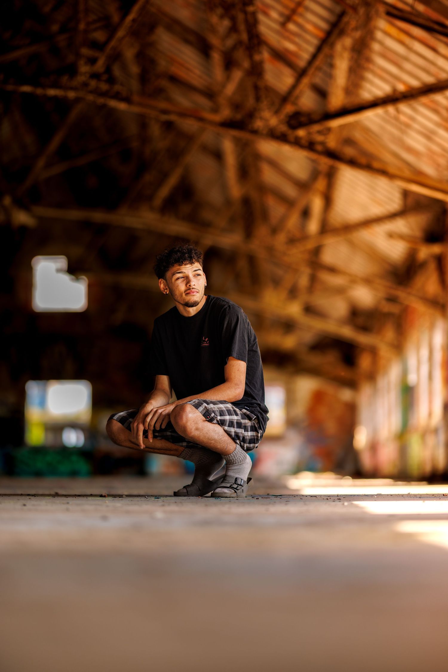 Young man in black t-shirt crouching in rustic wooden barn with warm sunlight streaming through windows.
