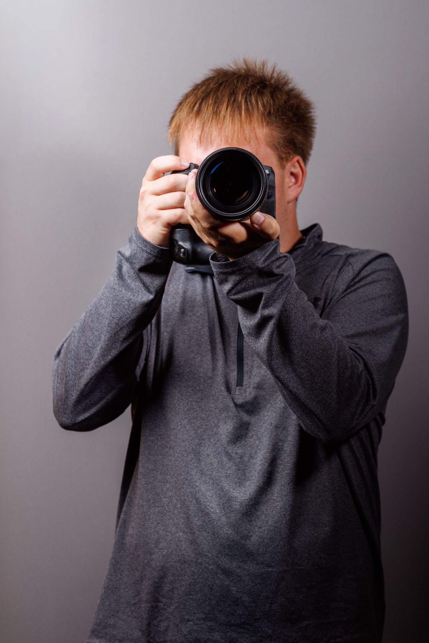 A young man with red hair holds a DSLR camera up to his face against a gray background.
