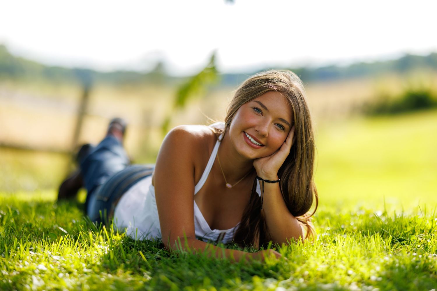 A smiling young woman with long brown hair lies on green grass in warm sunlight.