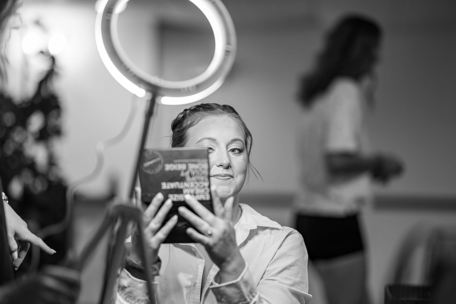 A bride examines her makeup palette under a ring light while preparing for her wedding day in black and white.