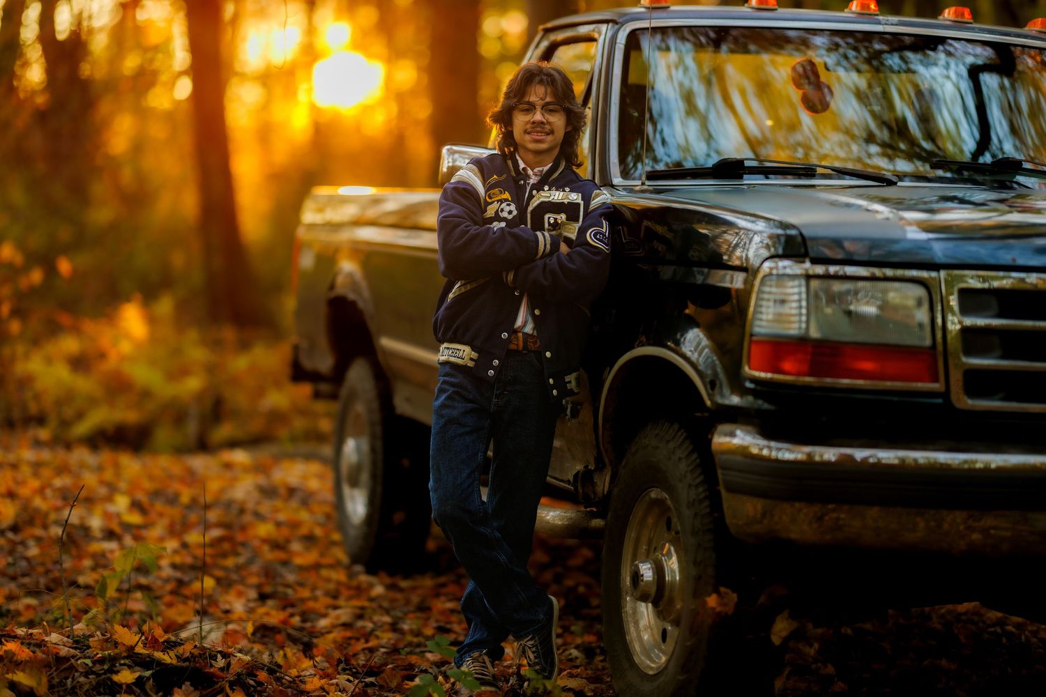 A young boy leans against a black pickup truck surrounded by autumn foliage at golden hour.