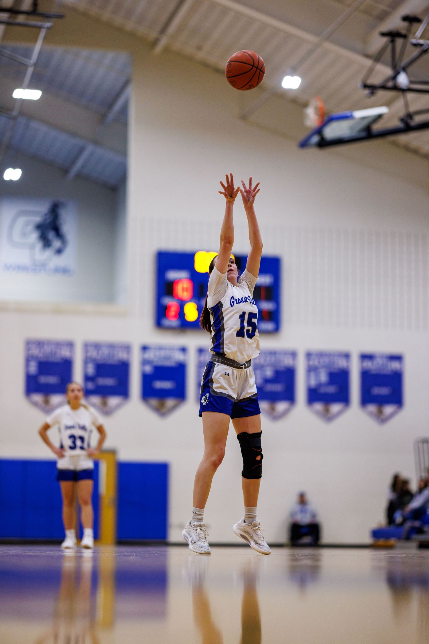 Female basketball player #15 shoots a jump shot during a high school girls basketball game in a blue and white gymnasium.