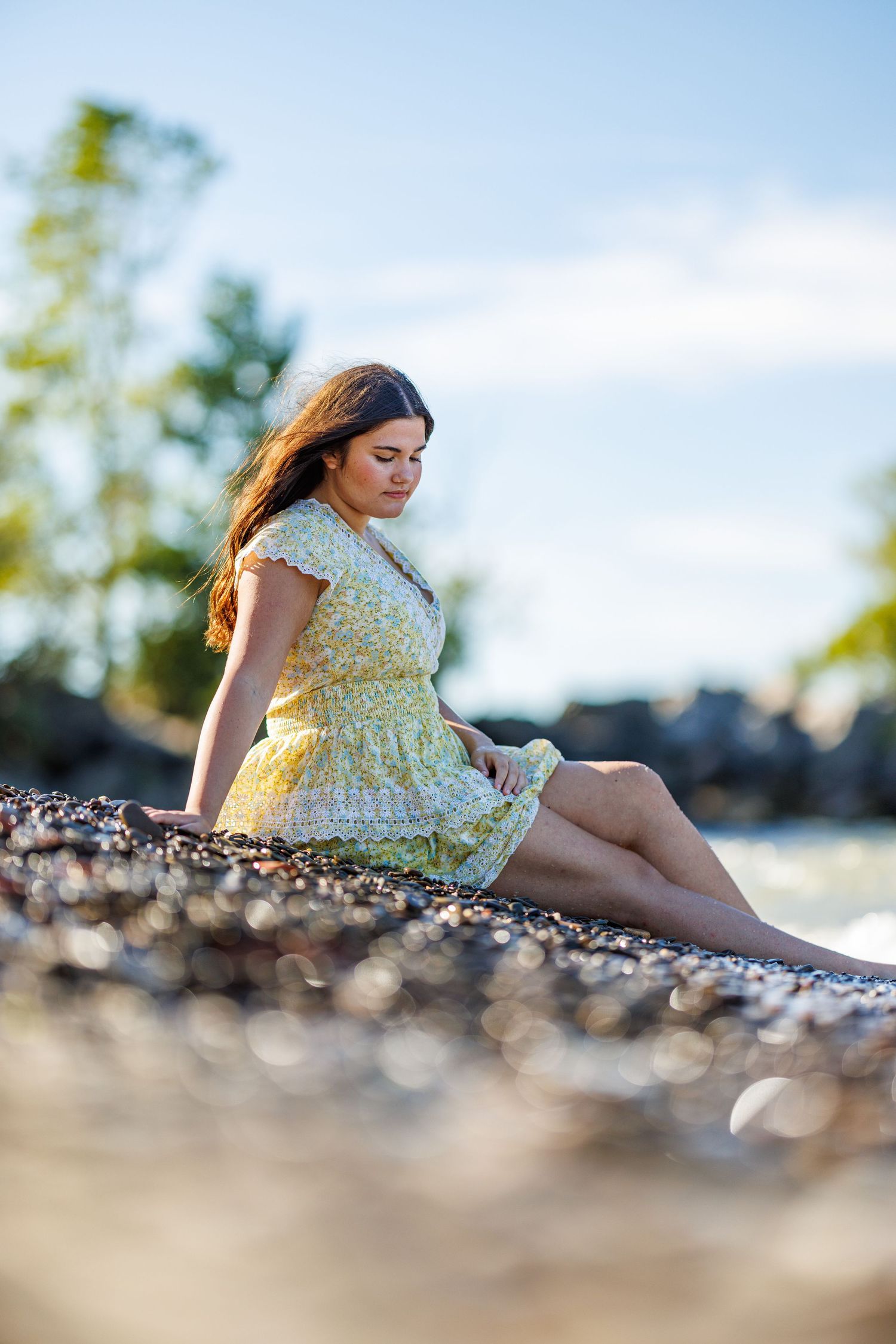 Young woman in yellow sundress sitting on pebble beach with wind in her hair.