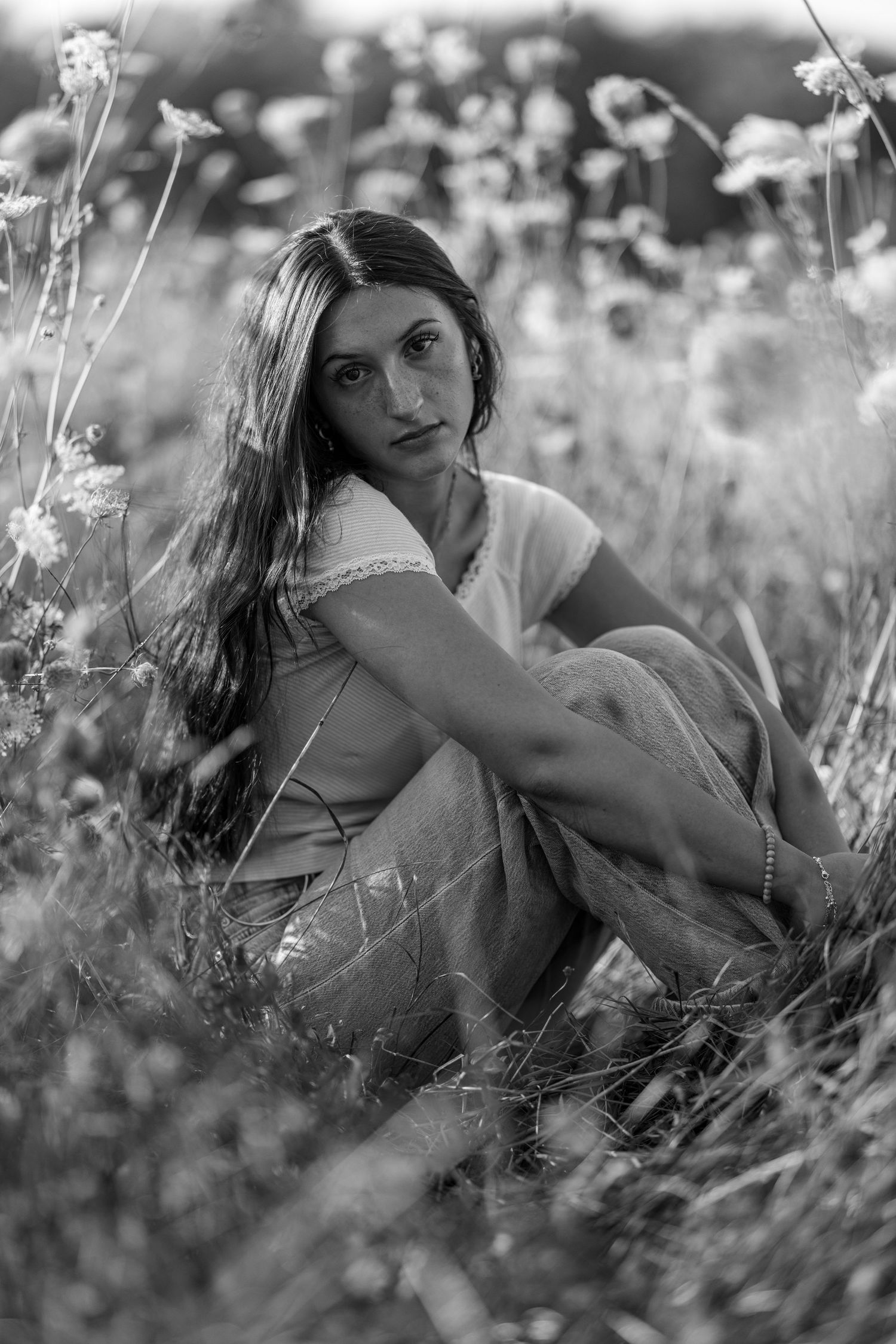 Black and white portrait of a young woman sitting cross-legged among wildflowers in a summer meadow.