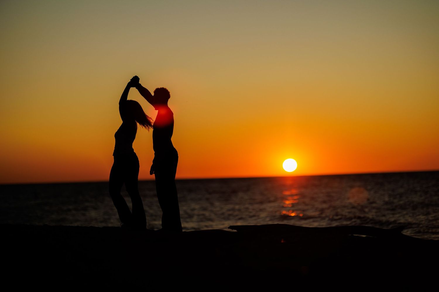 Romantic couple silhouetted dancing on beach at vibrant orange sunset.