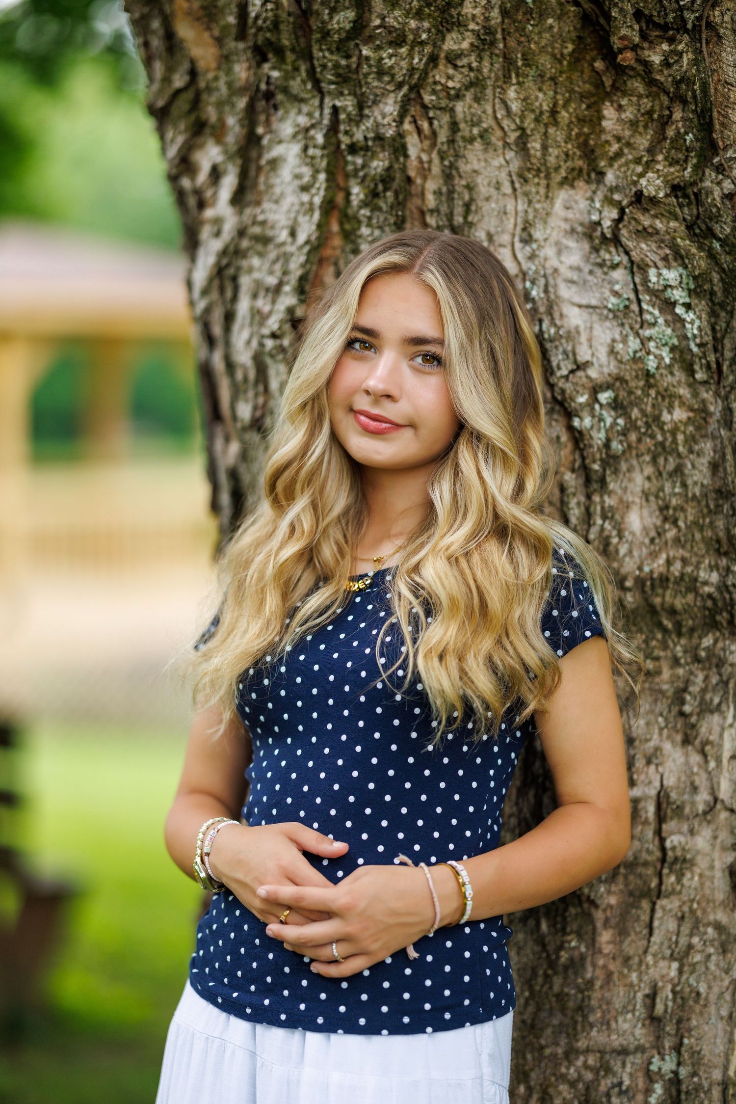 A teenage girl with long blonde wavy hair leans against a tree wearing a navy polka dot top.