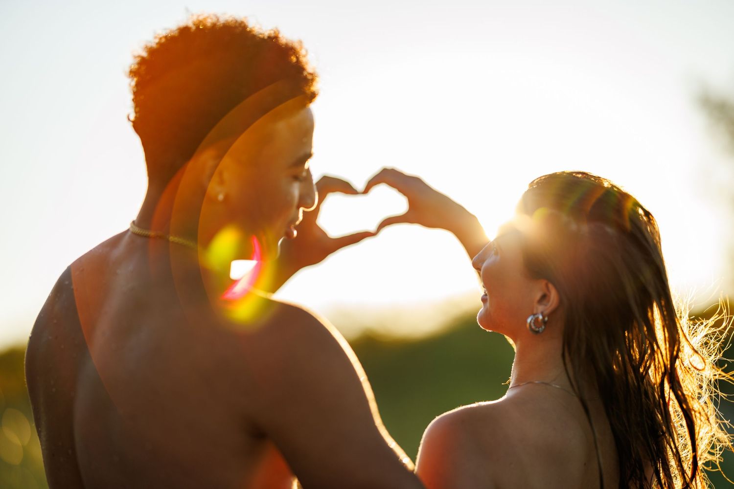 A couple making a heart shape with their hands at golden sunset, facing each other romantically.