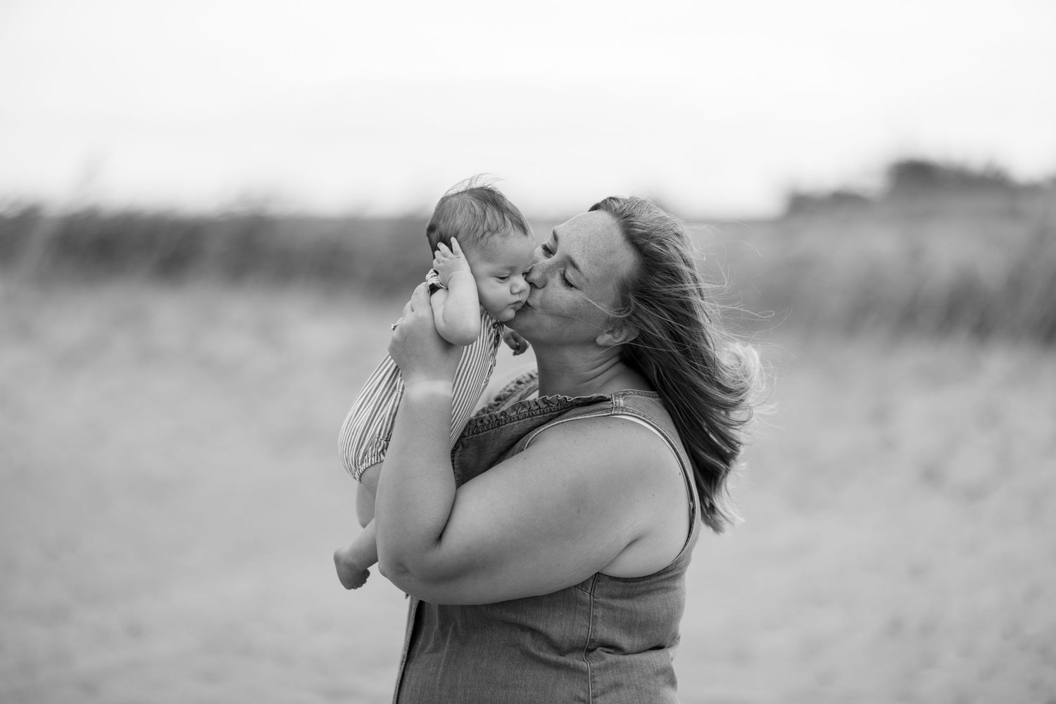 A mother tenderly kisses her baby outdoors in a beautiful black and white portrait.