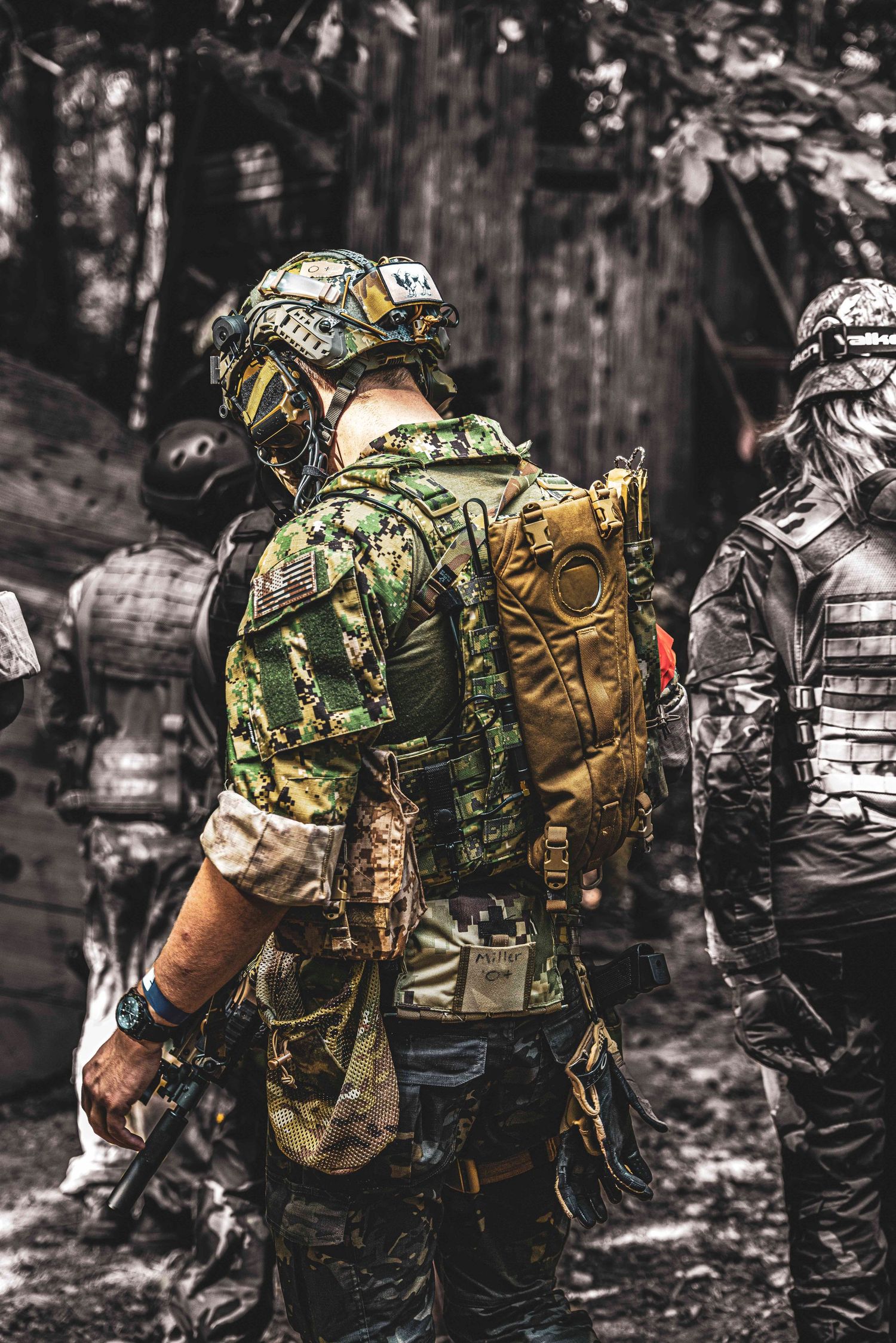 Fully geared airsoft players in military camouflage gear stand in a forest during a game.