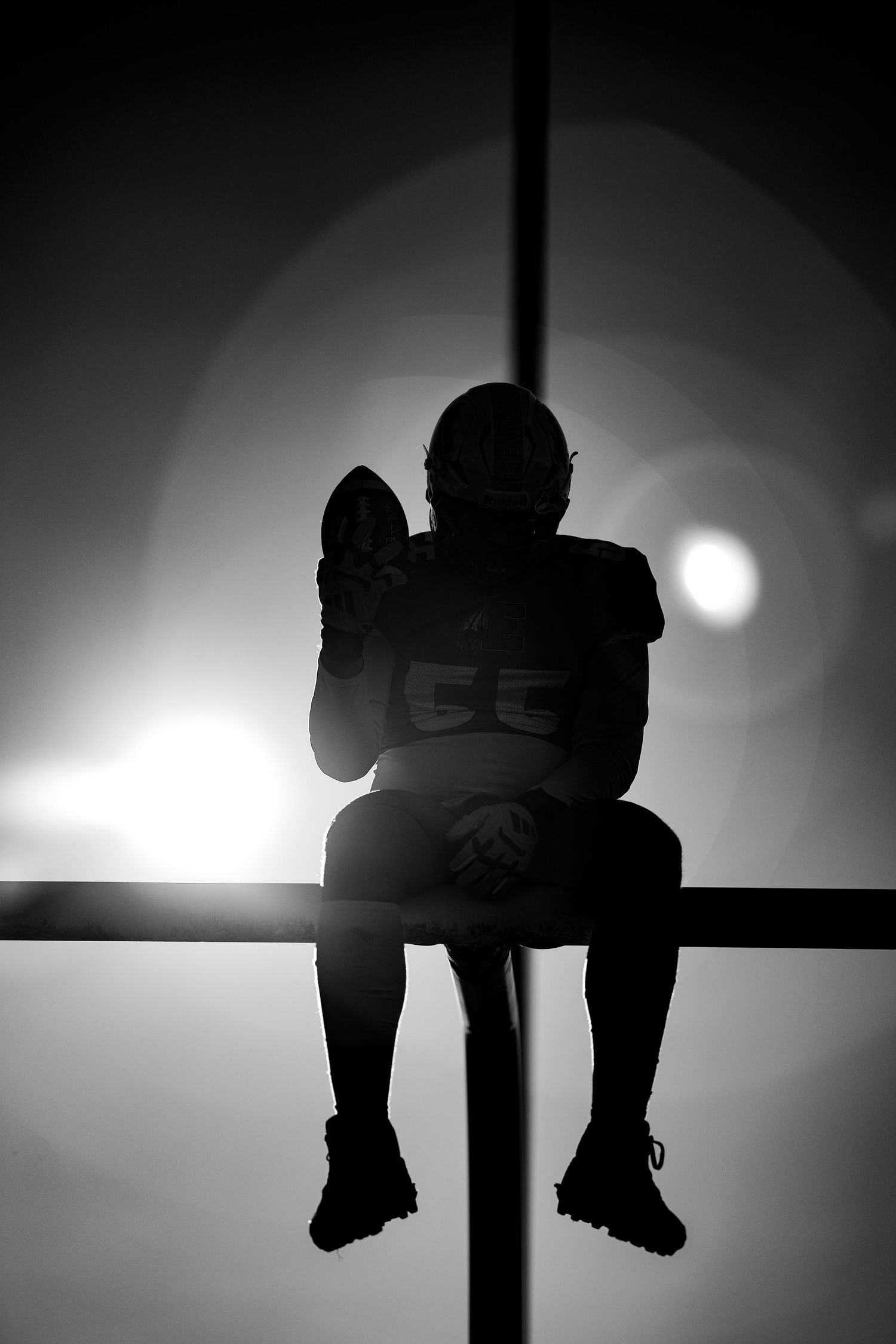 Silhouette of football player number 55 sitting on railing holding helmet against bright stadium lights.