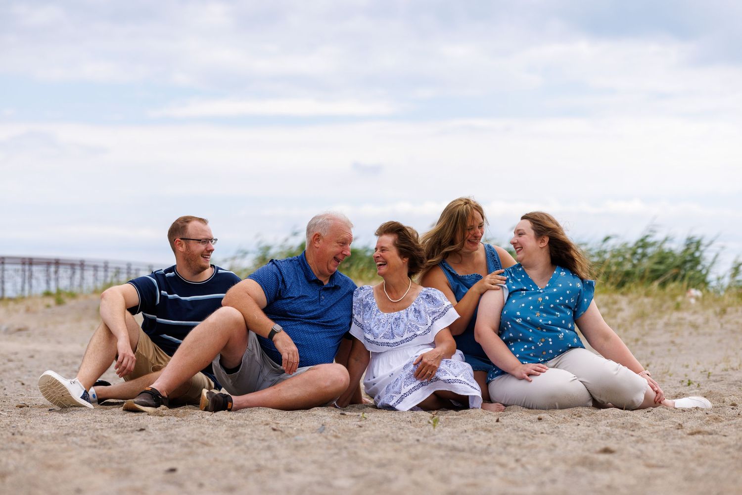 A multigenerational family sits together laughing on a sandy beach under a cloudy sky.