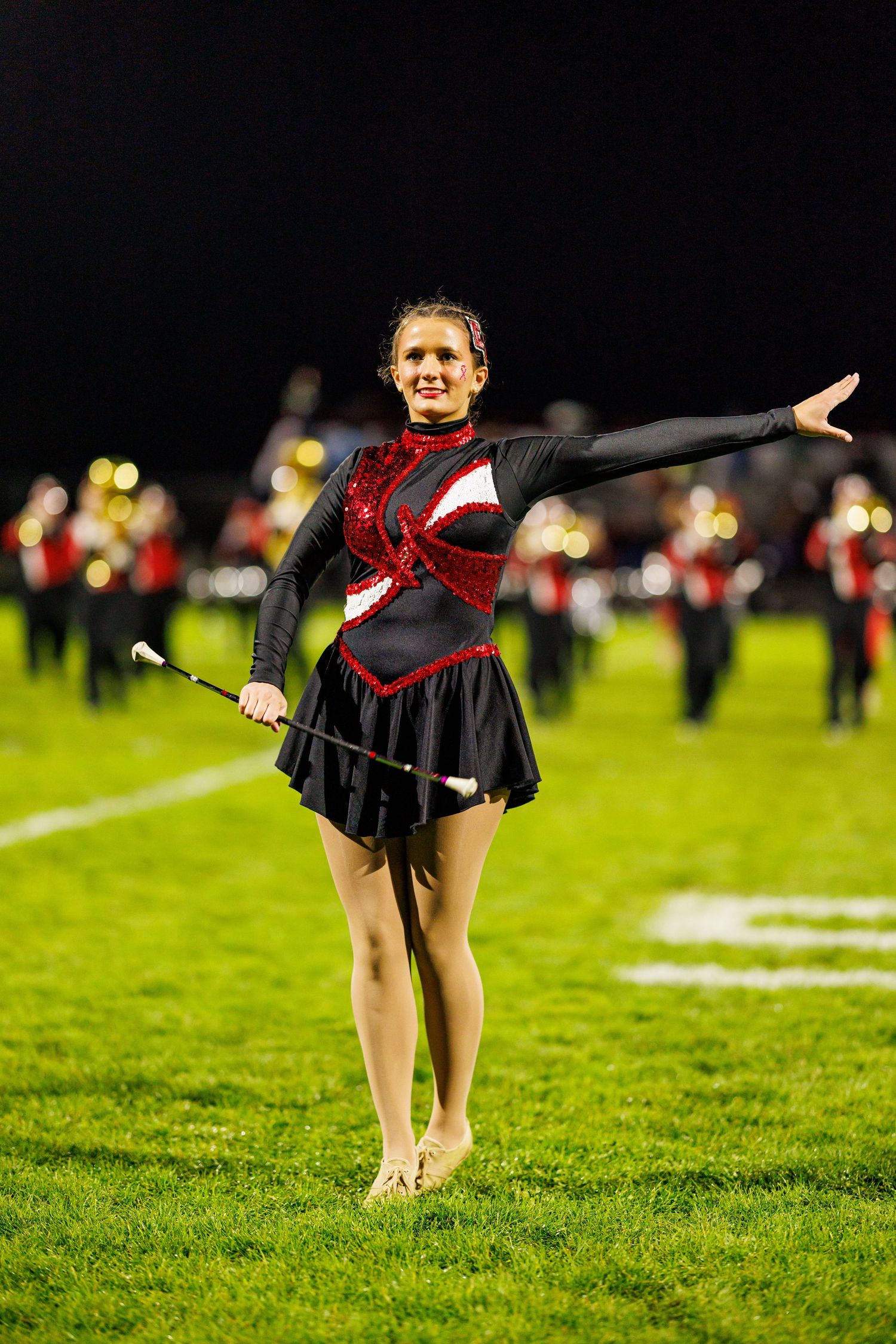 A smiling majorette in a red and black sequined uniform performs with a baton on a football field at night.